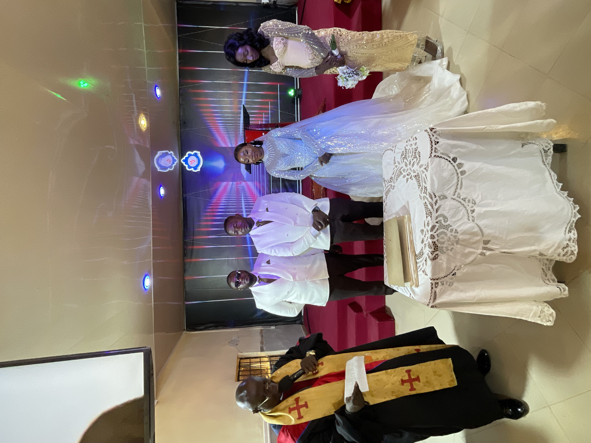 The image captures a wedding ceremony. A priest stands in the foreground, wearing a black robe with a gold and red stole adorned with crosses, holding a microphone and a piece of paper, likely performing the ceremony. Behind him are two men dressed in white suits and dark pants, presumably the groom and his best man. Further back, the bride is positioned in the center, wearing a shimmering white dress, and next to her is another woman in a gold dress, possibly the maid of honor. The ceremony is taking place on a red carpet, with a table draped in a white lace cloth to the right. The background features a screen with illuminated designs. The location appears to be a church or religious venue in the city of Unknown, Unknown.