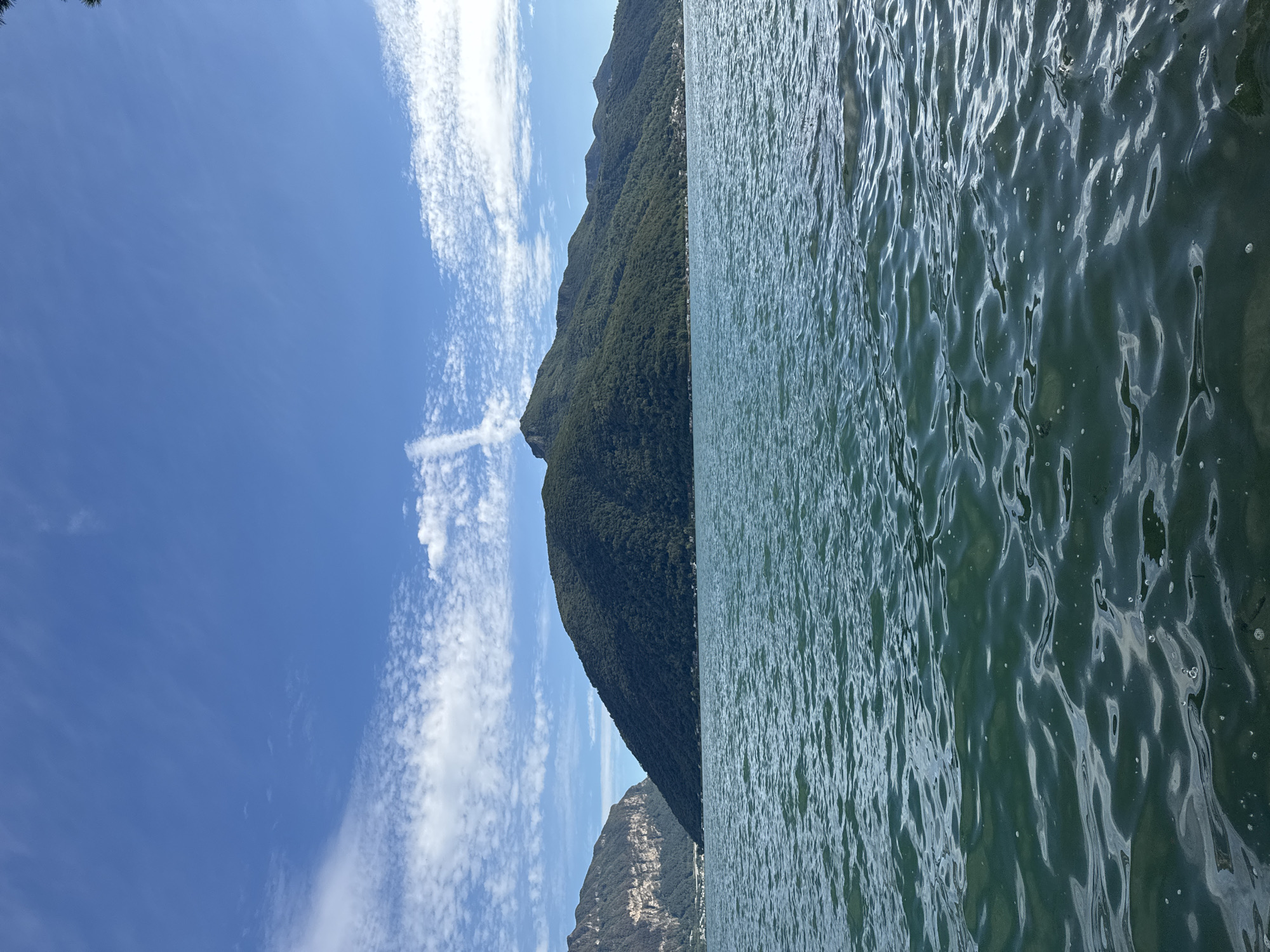 A wide shot captures a serene lake with a mountain range in the background under a bright blue sky with scattered white clouds. The foreground is dominated by the calm, green waters of Lake Lugano, with subtle ripples reflecting the light. In the mid-ground, a large, tree-covered mountain rises from the water, its slopes dotted with small buildings, indicating a settlement along the shore. To the left of this prominent peak, another, steeper mountain with rocky cliffs is visible. The sky above is a vibrant blue, featuring wispy cirrus clouds and a distinct contrail forming a cross shape. The overall scene suggests a peaceful, natural landscape in the vicinity of Melide, Switzerland, likely viewed from a boat or the shoreline.