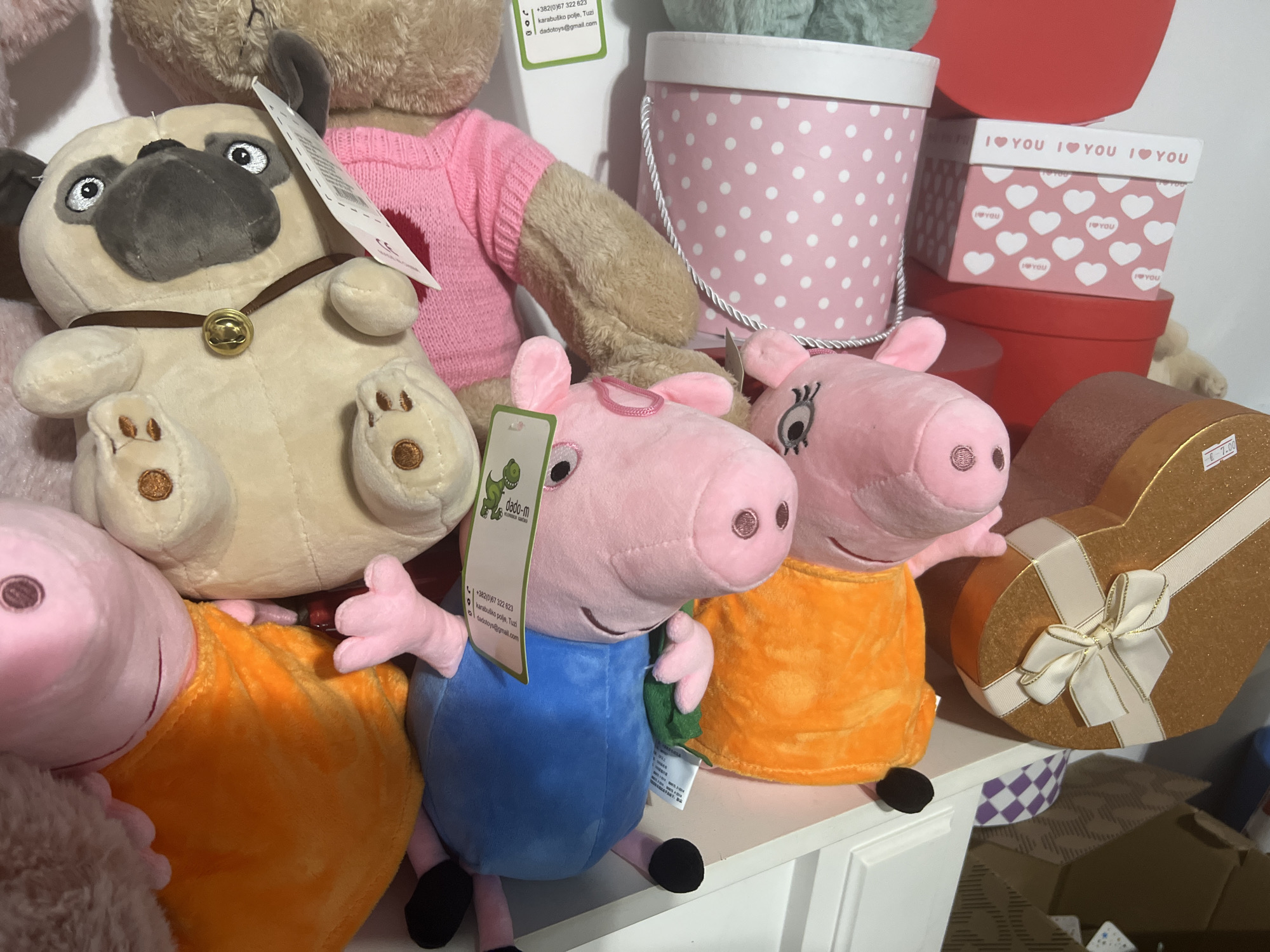 The image presents a collection of stuffed animals and gift boxes displayed on a white shelf. Featured prominently are two plush figures of Peppa Pig, one in a blue outfit and the other in orange, along with a stuffed pug dog. Adjacent to the toys are several gift boxes, including a white polka-dotted box, a red box, and a heart-shaped box adorned with a ribbon. The scene is well-lit and appears to be inside a retail establishment. Location mentioned on tags: Tuzi, Montenegro.