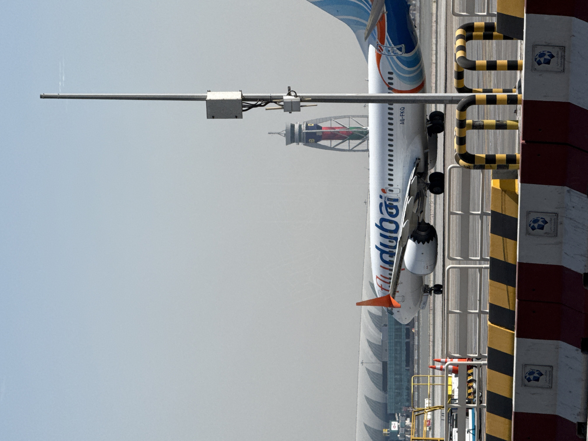 The image depicts an airplane with "flydubai" branding on the tarmac at Dubai International Airport (DXB). The aircraft is a Boeing 737, as indicated by the registration number A6-FKQ visible on its fuselage. In the background, the distinctive architecture of Dubai International Airport is visible, including a long, curved terminal building with a wave-like roof structure. A control tower with a unique design, featuring a cylindrical observation deck and a metal lattice structure, stands prominently in the middle ground. Several barriers, including those with black and yellow hazard stripes and concrete barriers with red and white markings, are positioned in the foreground, suggesting a secure operational area. The sky is clear and hazy, typical of the desert climate. No people are clearly visible in the image.