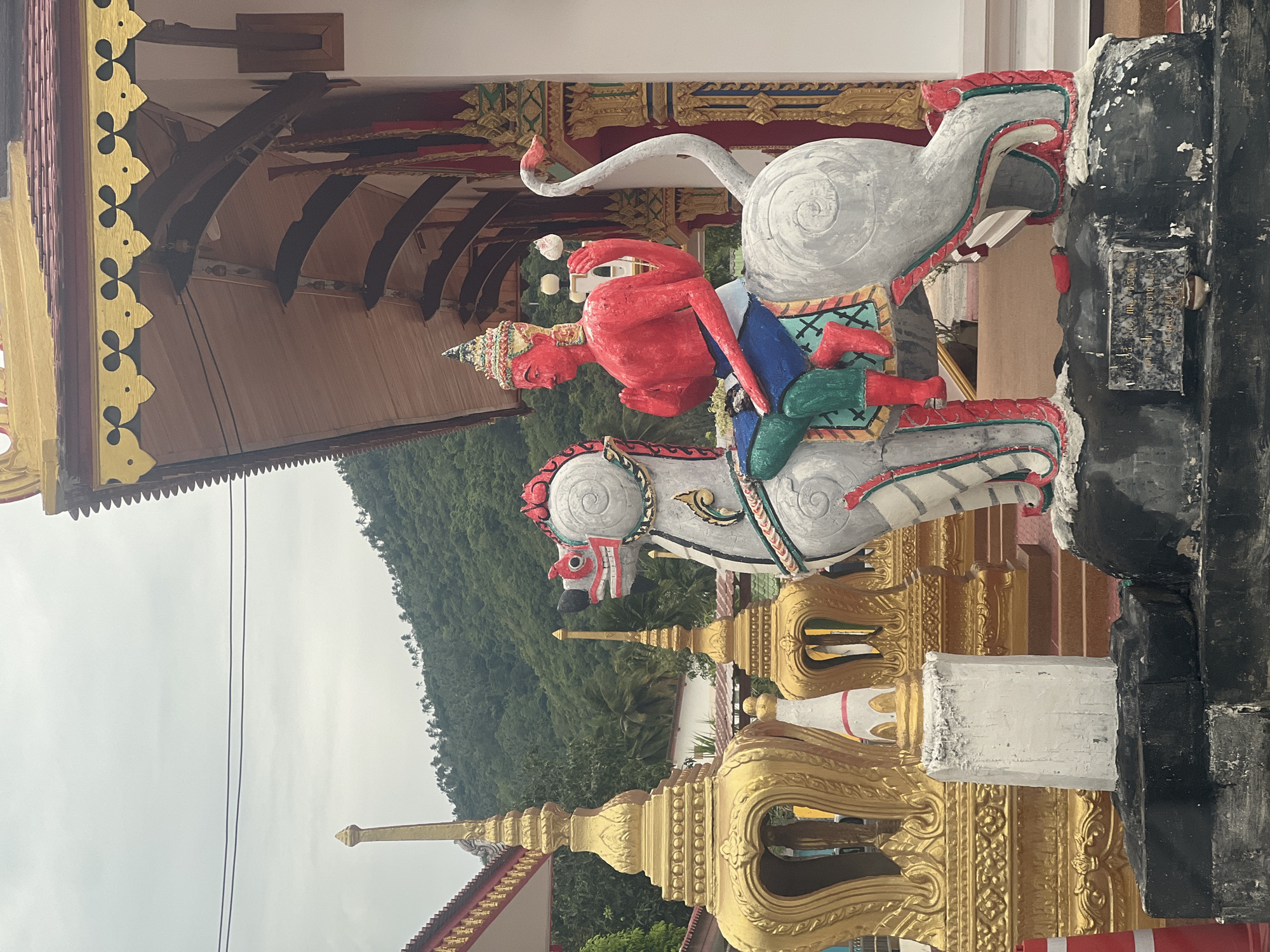 The image shows a brightly colored statue of a four-armed, red-skinned humanoid figure riding a mythical creature resembling a horse with a swan-like neck and tail. The statue is adorned with intricate patterns and appears to be made of concrete or stucco, with painted details in red, white, blue, and green. The figure wears a jeweled headdress and a blue garment around its waist, with green and red leggings. The mythical creature is predominantly white and gray, with red accents around its eyes and mane.
The statue is positioned in front of a building with a traditional Southeast Asian architectural style, featuring a sloped, tiled roof with ornate golden trim and decorative carvings. To the left of the statue, there are several golden, tiered structures, possibly stupas or decorative pillars, also characteristic of Buddhist temple architecture. In the background, a lush, green, tree-covered hillside rises under a cloudy sky. The overall scene suggests a religious or cultural site, likely a Buddhist temple.