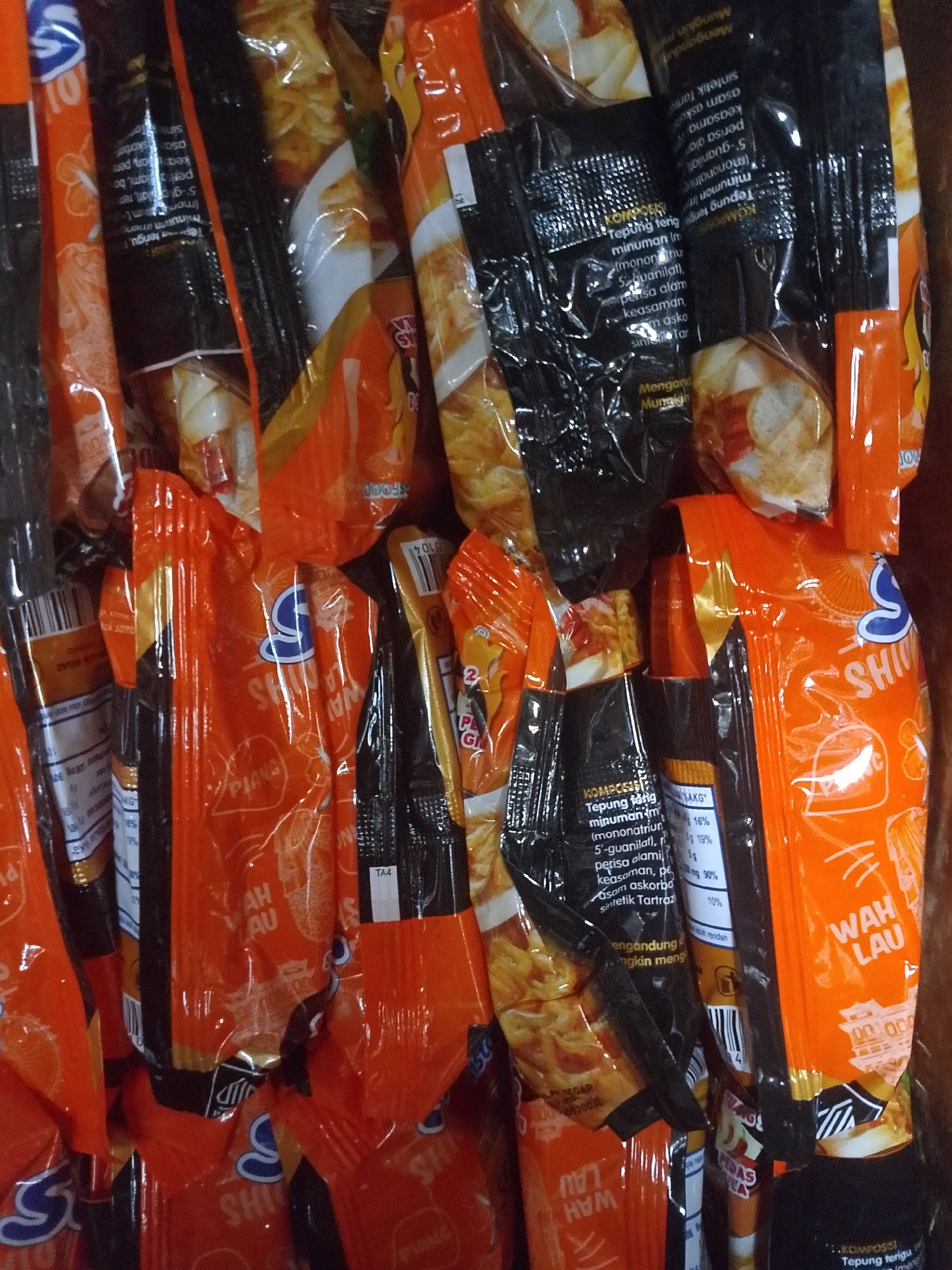 The image displays a close-up, slightly blurred, top-down view of a stack of packaged instant noodles. The packaging is predominantly orange and black, with some white and blue accents. The text on the packaging is in a foreign language, likely Indonesian, given the mention of Mukapayung, Indonesia. Several packages have graphics of stylized ramen bowls or noodle dishes. The lighting appears to be ambient, with some reflections on the plastic packaging, suggesting an indoor setting. The overall impression is of a commercial display or storage of food products.