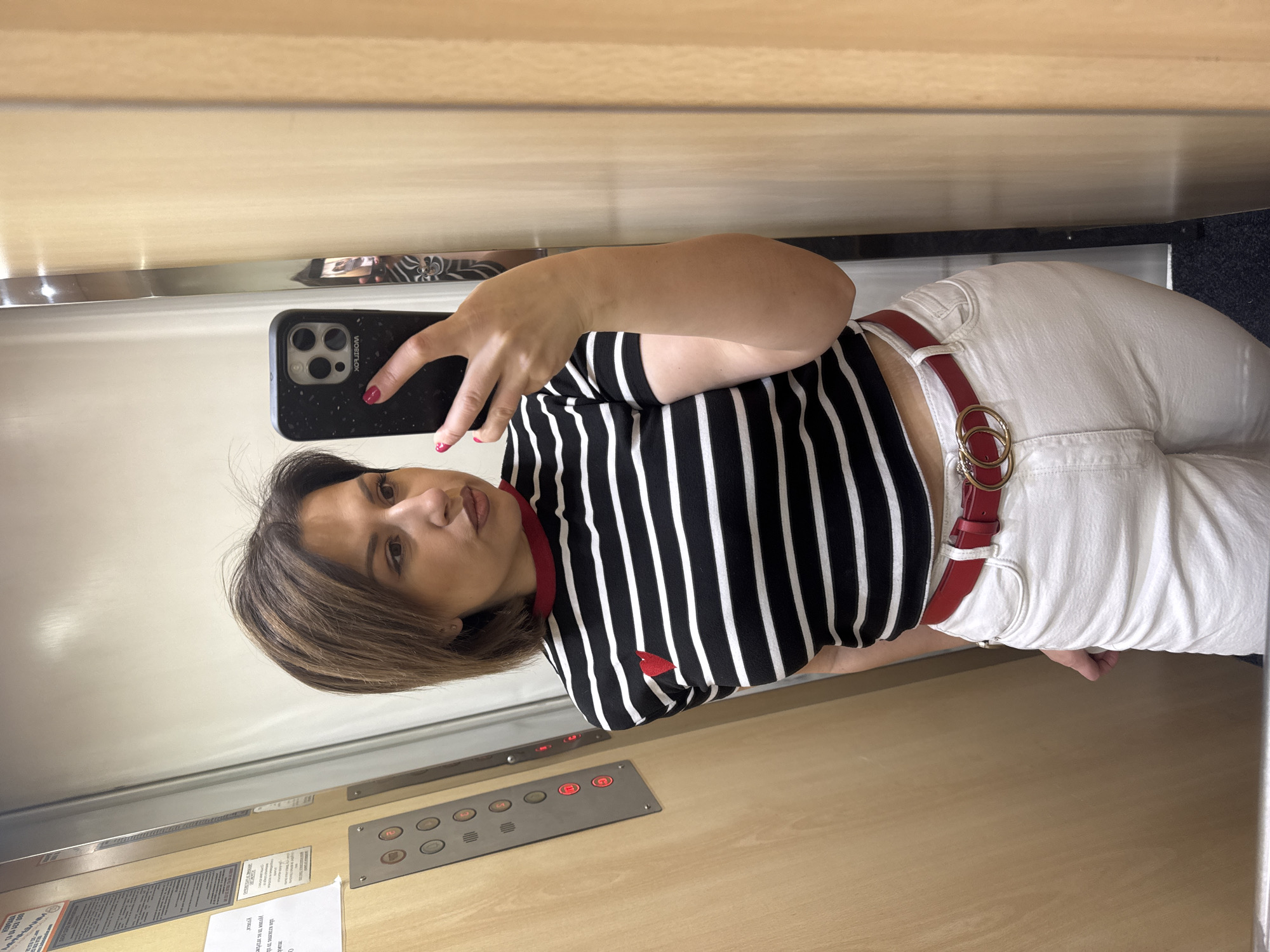 A woman is taking a selfie in an elevator. She is wearing a black and white striped shirt with a red collar and a small red heart emblem, white pants with a red belt featuring a double-ring buckle, and is holding a smartphone with a black case. The elevator interior consists of wood-paneled walls with a control panel visible containing several buttons. Based on the elevator controls written in Bulgarian, the elevator is likely in Sofia, Bulgaria. The lighting is bright and even, suggesting fluorescent or LED fixtures.