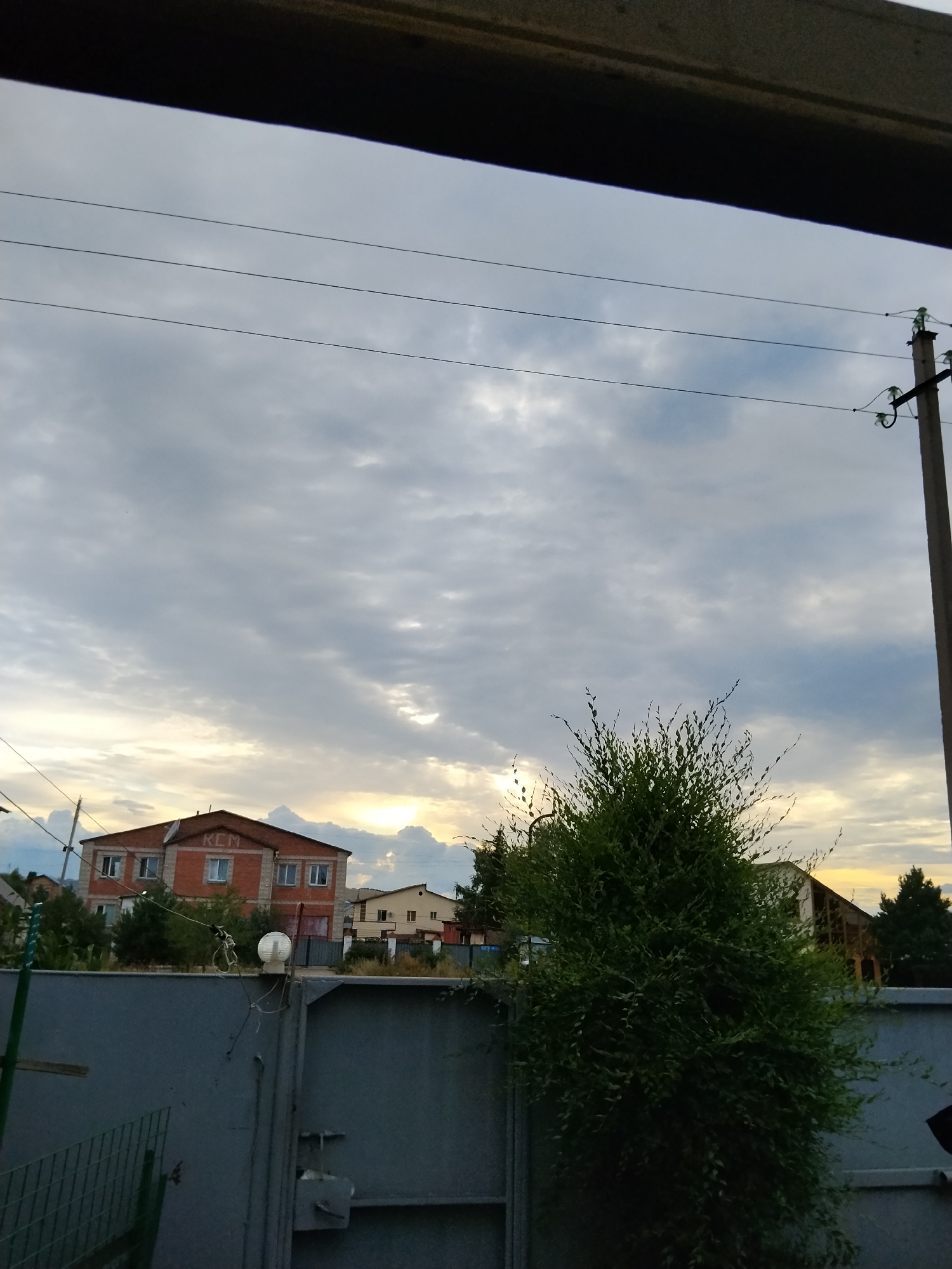 The image depicts a cloudy sky over a residential area, likely at dusk. A large, multi-story brick building with multiple windows is visible on the left side, and a smaller, lighter-colored building is to its right. In the foreground, a tall, dense green bush or small tree stands in front of a grey metal fence. Overhead, power lines stretch across the frame, supported by a concrete utility pole on the right. The sky is a mix of grey clouds with patches of lighter sky and sunlight breaking through in the distance. The scene appears to be an outdoor, semi-urban or suburban setting.