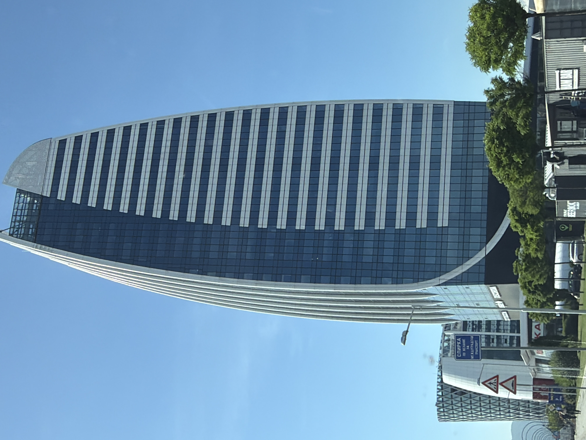 The image shows a modern skyscraper with a curved, white facade and a blue glass exterior in Sofia, Bulgaria. The skyscraper is tall and slender, with horizontal white bands across its facade. In the foreground, there are some trees, street signs, and a few people walking. In the background, other buildings and structures can be seen. The sky is clear and blue. The signs in the image are in Bulgarian. One sign reads "СПИРКА" which means "STOP". Another sign indicates a pedestrian crossing. The skyscraper appears to be a prominent architectural landmark in the city. The overall scene suggests an urban environment on a sunny day.