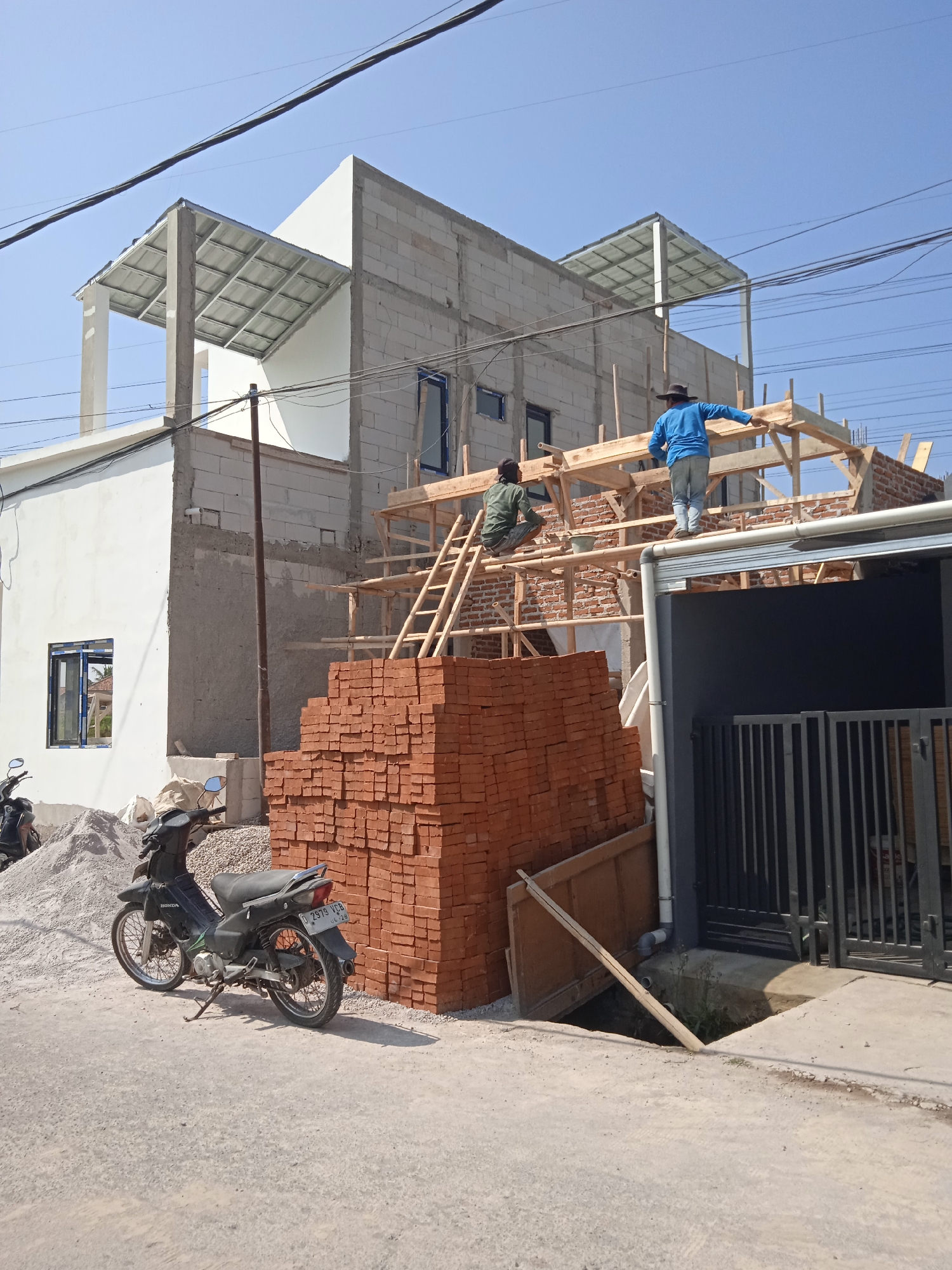 This image captures a construction site in Cileunyi, Indonesia, where a multi-story building is under construction. The building's exterior walls appear to be made of concrete blocks, with some sections already plastered. Two construction workers are visible on the partially constructed wooden framework of the upper floor. One worker, wearing a blue shirt and a hat, stands and is adjusting a wooden beam. The other worker, in a green shirt and hat, is seated on a wooden ladder. In the foreground, a large stack of red bricks is piled high, indicating materials ready for use. A black motorcycle is parked on the gravelly ground next to the brick pile, with a pile of sand also visible. The scene is set against a clear blue sky, with power lines crisscrossing overhead. To the right, a portion of a dark-colored fence with vertical bars is visible, suggesting the presence of an adjacent property. The ground surface is a mixture of concrete and loose gravel.