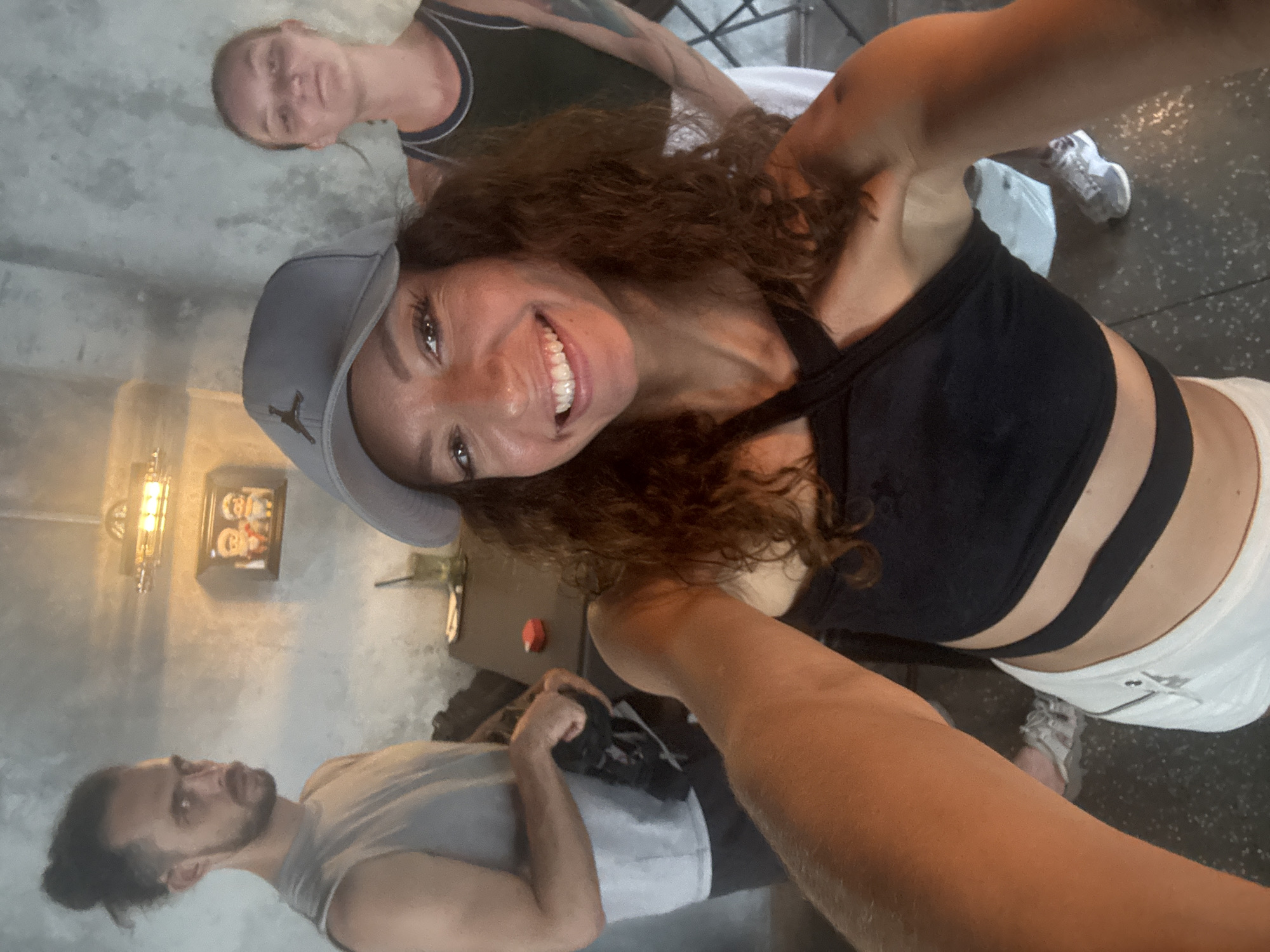 The image shows a selfie taken by a woman with curly brown hair, wearing a grey baseball cap with a Jumpman logo, and a black sports bra with white shorts. She is smiling at the camera. Behind her, to the left, is a man with a beard and a bun, wearing a light grey tank top. Behind the woman taking the selfie and slightly above is a woman with short hair, wearing a dark tank top with black straps. The background includes a grey wall with a framed picture and a decorative wall lamp. The floor appears to be dark grey. It is not possible to definitively determine the location as Đà Nẵng, Vietnam based on the visual information alone. There are no recognizable landmarks or environmental factors that would allow confirmation of the location.