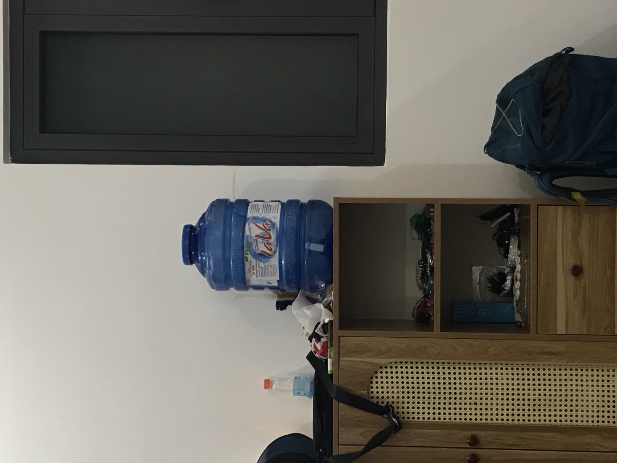 The image displays an indoor scene, likely a room in Mũi Né, Vietnam, featuring a white wall as the backdrop. High on the wall, a dark gray rectangular object, possibly a window frame or a wall-mounted screen, is visible.
Below this, a large, translucent blue 5-gallon water cooler bottle, emblazoned with the brand "La Vie," is positioned horizontally, resting on a surface next to a wooden cabinet. To the right of the water bottle, a wooden cabinet or shelf unit is prominent. The upper section of this unit consists of open shelves containing various small items and a closed wooden door with a round brown knob. Below this, a larger compartment features a door with a distinctive woven rattan-like pattern. A lower drawer with another brown knob is partially visible at the bottom of the frame.
A blue backpack with yellow elastic cording is seen on the upper right side of the image, appearing to rest on or near the top of the cabinet, partially against the white wall. Closer to the bottom left, a smaller, clear plastic water bottle can be seen amidst other miscellaneous items. The scene suggests a functional and lived-in space with everyday objects. No specific activity is taking place.