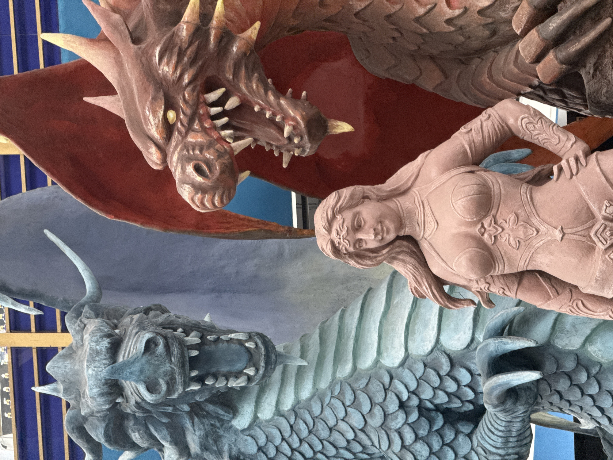 The image shows a detailed sculpture featuring two dragons and a female figure. One dragon is depicted in brown with yellow spikes and an open mouth revealing teeth. The other dragon is blue-gray with scales and also has an open mouth with visible teeth. A woman's sculpted figure lies between the two dragons, facing the viewer. The sculpture has an intricate level of detail.
The sculpture is located in Nakhodka, Russia.