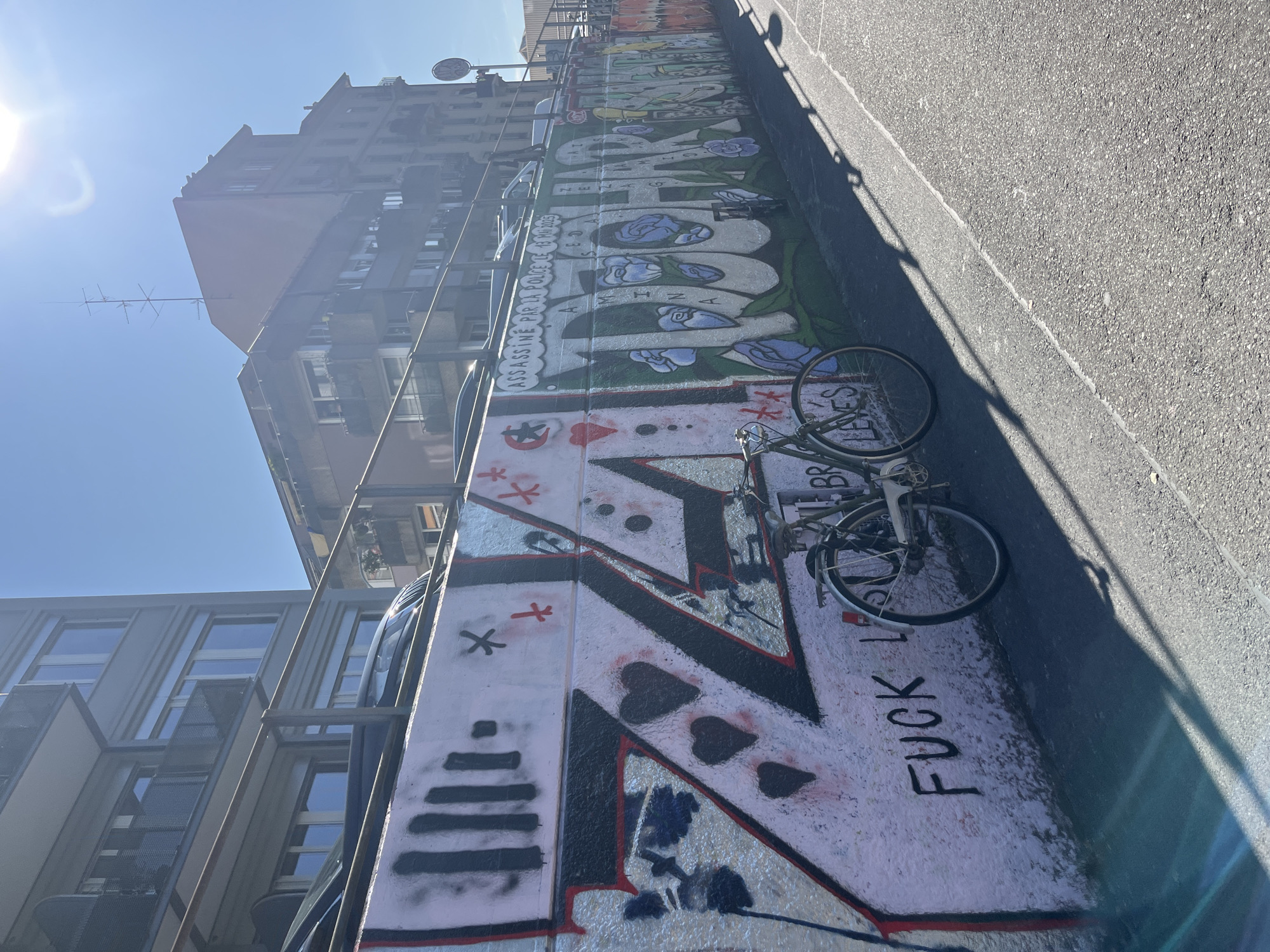 An outdoor shot shows a graffiti-covered wall next to a road in Geneva, Switzerland. The graffiti features large letters, hearts, and flowers. A bicycle is parked against the wall. In the background, buildings and a clear blue sky are visible. The sun is shining brightly, casting shadows on the road. The graffiti appears to be a political statement, with the phrase "ASSASSINE PAR LA POLICE LE 13 MAI 2023" visible on the wall. The overall scene depicts an urban environment with elements of street art and protest.