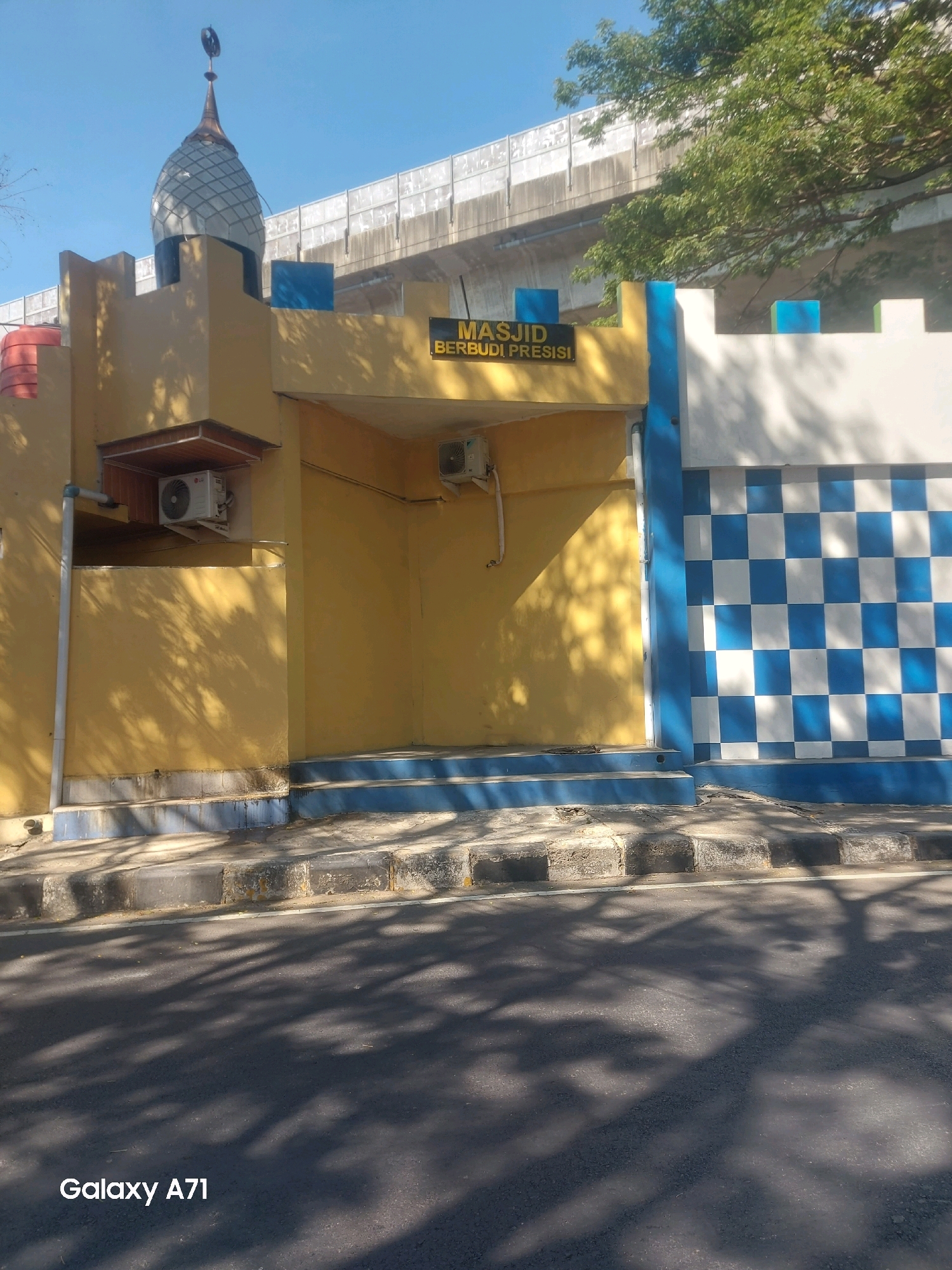The image shows the exterior of a mosque with a yellow facade and a blue and white checkered wall. A sign above the entrance reads "MASJID BERBUDI PRESISI". A dome with a crescent moon topping is visible on the left side of the building. An elevated road or bridge is visible in the background, partially obscured by trees. The foreground shows a street with shadows from trees cast upon it. The bottom left corner of the image contains the text "Galaxy A71". This mosque is located on Jalan Ratulangi, in the city of Makassar, Indonesia.