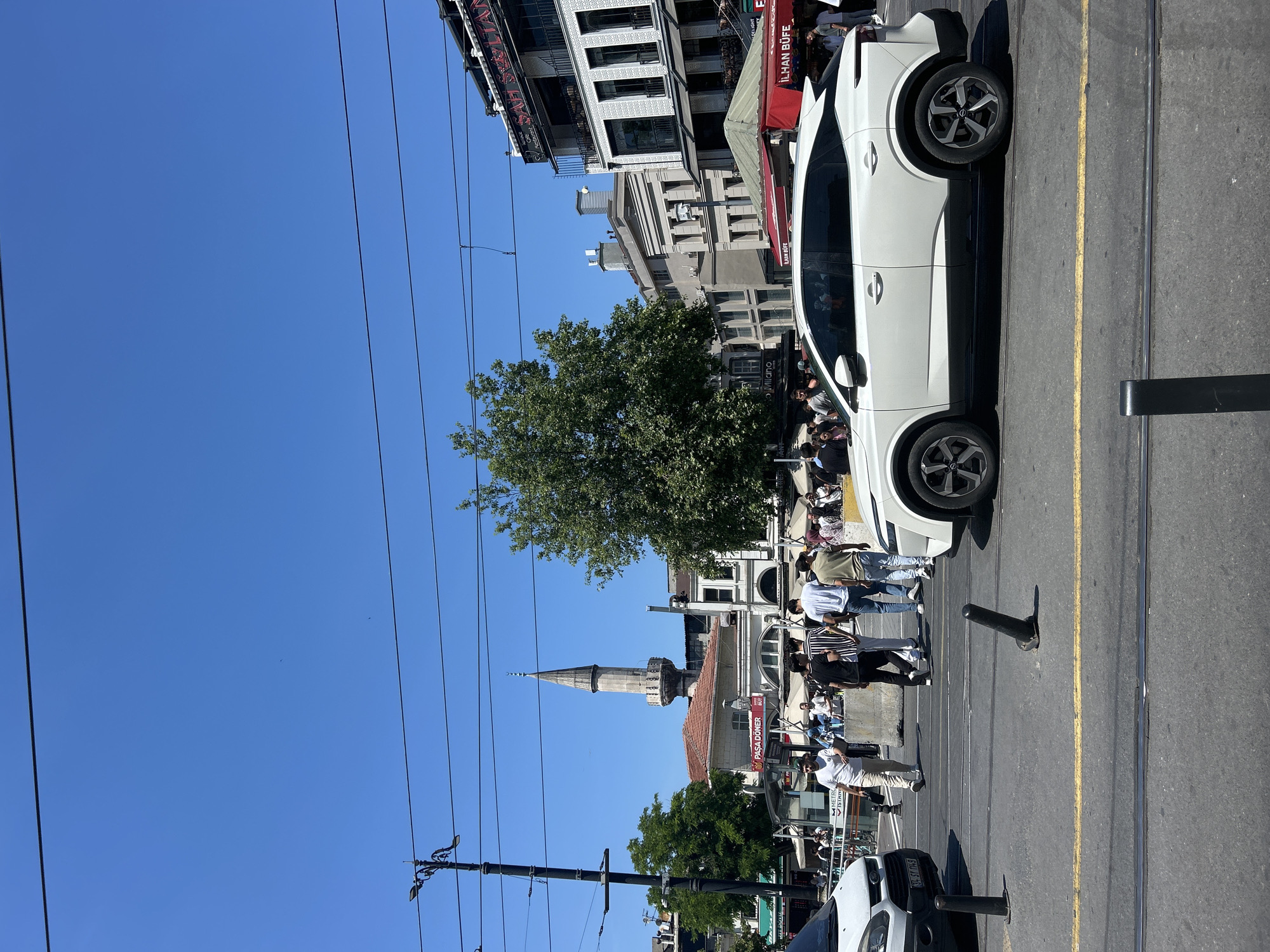 The image depicts a street scene in Istanbul, Turkey, likely in a bustling urban area given the presence of a tram track, overhead wires, and a crowd of people. A white SUV is the dominant subject, positioned in the foreground and slightly to the right, facing left. Behind the SUV, several pedestrians are walking across a street intersection.
In the background, a prominent minaret of a mosque rises against a clear blue sky. Adjacent to the mosque, buildings with architectural styles suggestive of historical and modern influences are visible. Some of these buildings appear to house businesses, with awnings and signage indicating food establishments like "PAŞA DÖNER" and "İLHAN BUFE." Trees, including a large, leafy one near the center of the frame, provide greenery.
The environment is characterized by a bright, sunny day, with a distinct lack of cloud cover. The street is paved, and tram tracks are clearly embedded in the asphalt. Several dark, bollard-like posts are present on the road, some appearing to be traffic control devices. The overall impression is that of a lively public space in Istanbul, capturing a moment of everyday life with a blend of historical and contemporary elements.