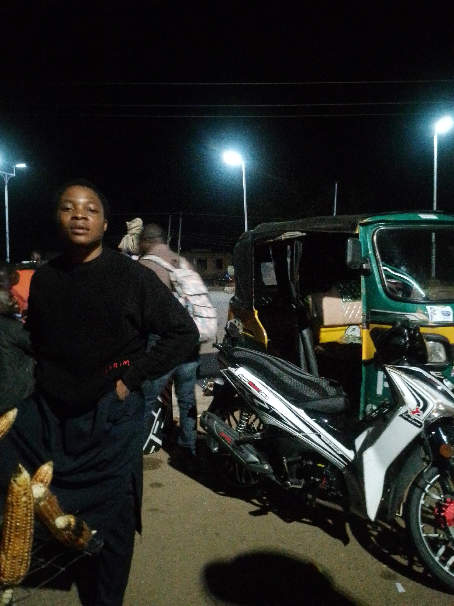 The image depicts an outdoor scene at night in Bukuru, Nigeria, illuminated by streetlights and the headlights of a motorcycle. In the foreground, a woman stands facing the camera, wearing a black long-sleeved shirt and dark trousers. To her left, two ears of corn are displayed on a rack. Behind the woman, a green and yellow auto-rickshaw is parked, with a white and black motorcycle in front of it. Other individuals are visible in the background, some carrying bags. The ground is paved, and electrical wires are strung overhead.