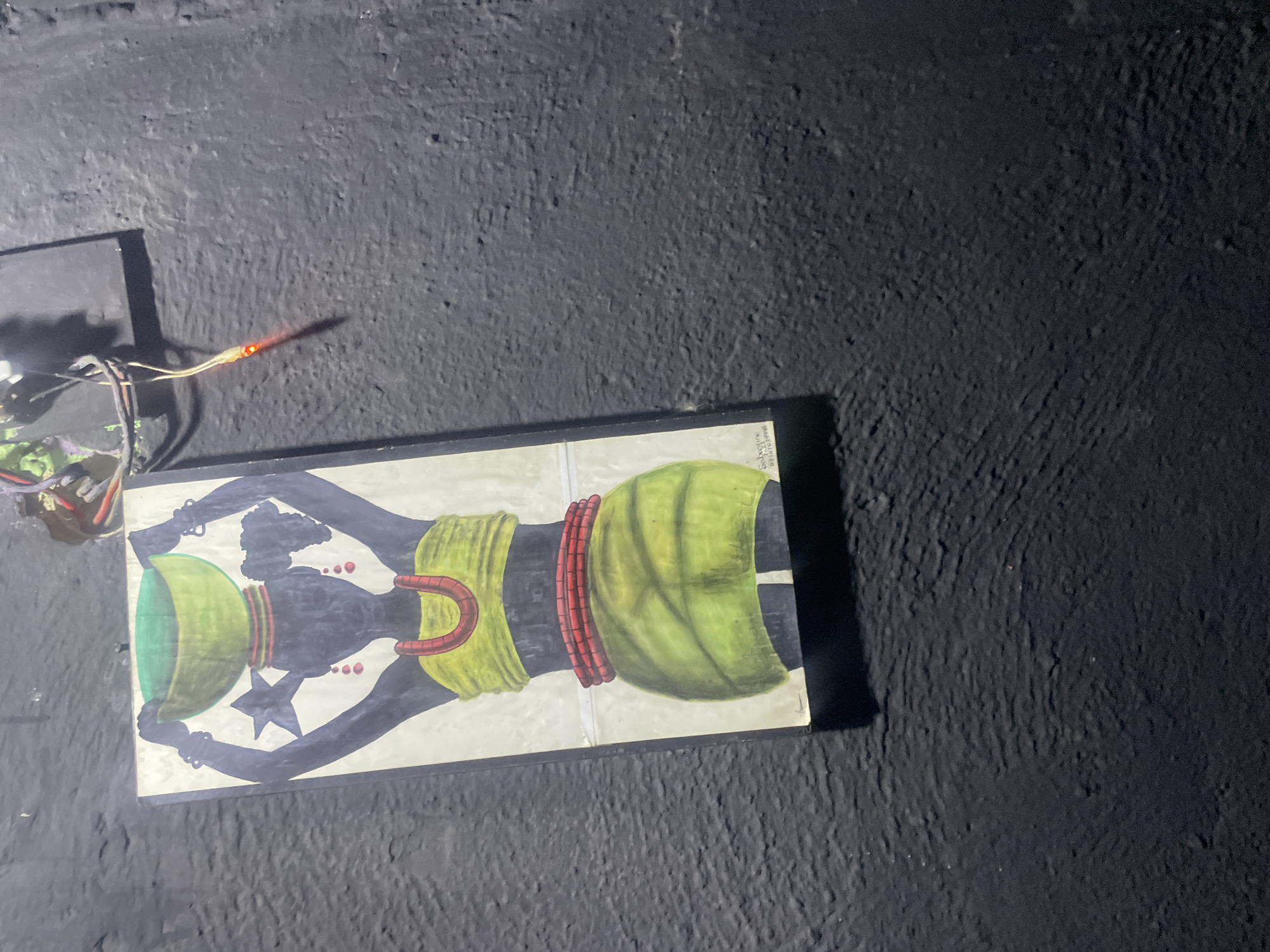 An illustration of a woman carrying a bowl on her head is taped to a textured black wall in Rafin Par, Nigeria. The woman in the illustration is silhouetted in dark blue, wearing a green top and skirt, and red jewelry. She has a star on her forehead. Wires and a red LED light are attached to the wall above the illustration.