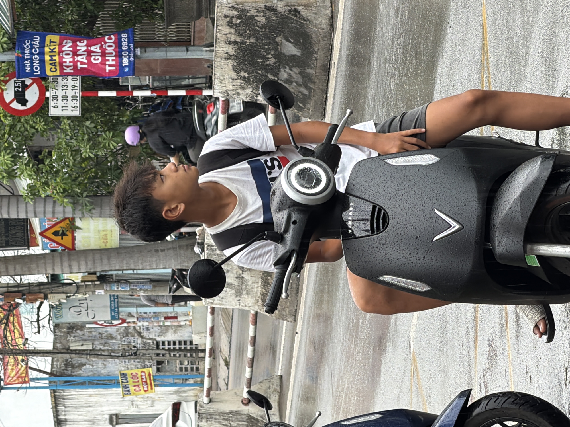 An outdoor medium shot captures a young male, possibly a teenager, seated on a black VinFast electric scooter in what appears to be a city street in Nha Trang, Vietnam. The scooter is dark gray and shows water droplets, indicating recent rain. The individual wears a white t-shirt with a navy and red stripe design across the chest and dark shorts. He has a black backpack on. His left foot is visible, wearing a patterned sandal.
The background is somewhat blurred but reveals a busy urban environment. To the left, a white car is partially visible. A concrete barrier with red and white stripes runs horizontally across the mid-ground. In the distance, there are buildings, trees, and various signs, typical of a commercial or residential area. One prominent sign on a pole to the right is a vertical banner with Vietnamese text, appearing to be an advertisement. Another sign above it is a circular red and white traffic sign with a black truck symbol and the number "2.5t". A white rectangular sign below it displays times in Vietnamese. A yellow sign with Vietnamese text is visible on the left. Palm trees are interspersed throughout the background, and streetlights and utility poles are also present. The ground is wet and reflective, suggesting it is raining or has recently rained. Another electric scooter, dark blue, is partially visible to the left of the frame. A woman with a purple helmet is seen riding another scooter in the background.