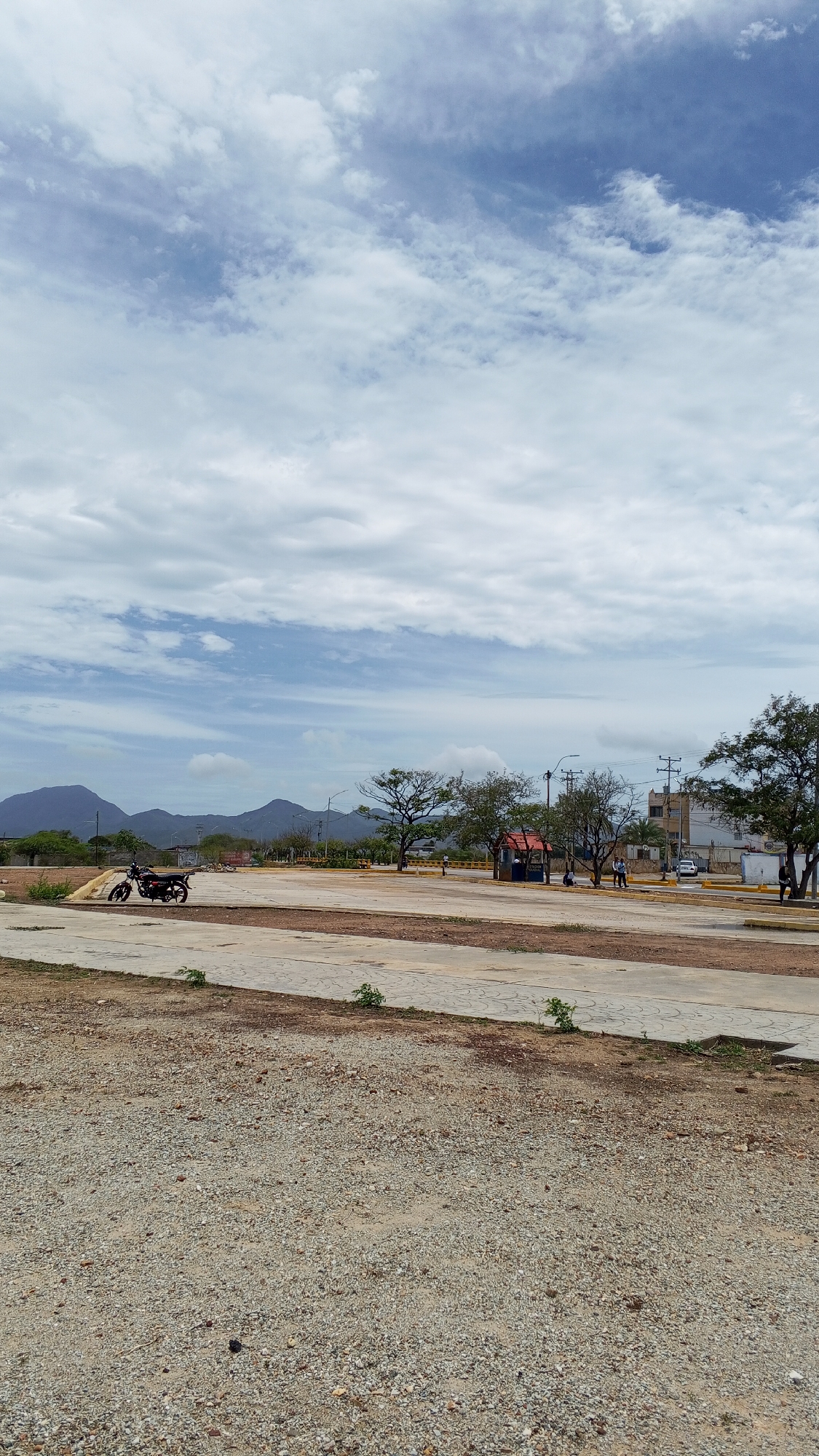 The image shows a wide-angle view of an open, partly-paved area in Juan Griego, Venezuela. The foreground consists of a gravel-covered surface. In the middle ground, there's a concrete area with various trees, a motorcycle parked to the left, a small red and blue structure resembling a bus stop. People stand nearby. In the background, are mountains and buildings under a partly cloudy sky. Utility poles and wires are also visible. The scene appears to be a large parking lot or open space in the town.