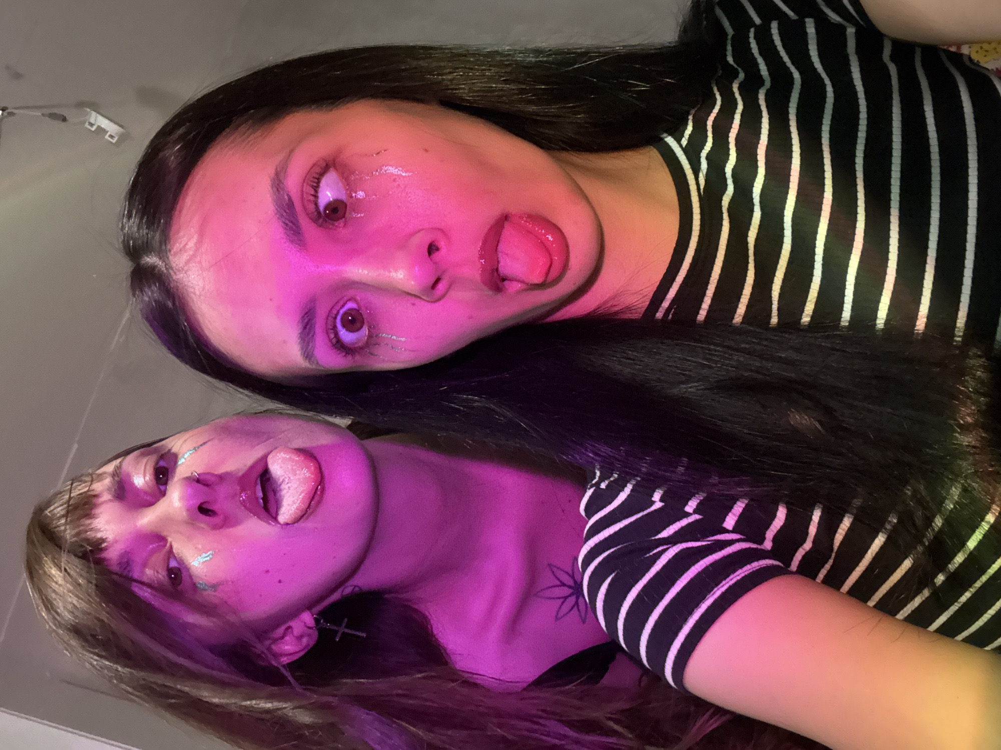 Two young women pose in a brightly lit, possibly indoor setting. Both stick their tongues out at the camera. The woman on top has long, dark hair and wears a black and white striped shirt. The woman on the bottom has shoulder-length brown hair with bangs and also wears a black and white striped shirt. Both appear to have some kind of glitter or makeup around their eyes. The scene is lit with a pink or purple light. There is no clear indication of the location being Votkinsk, Russia, or any other specific location.