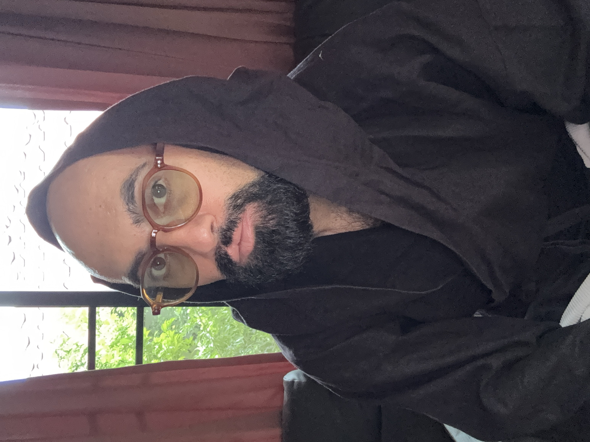 The image shows a medium close-up, head-and-shoulders portrait of a man wearing a dark hooded robe and round, amber-tinted glasses. He has a full beard and is looking directly at the camera. The background consists of a window with green foliage visible outside, and a reddish-brown curtain to the left and right of the window. The man appears to be indoors, seated on a dark couch. The lighting suggests an indoor setting, likely during daytime. The overall impression is a casual, slightly posed portrait. The specific location in Ko Samui District, Thailand, cannot be determined from the visual information provided.