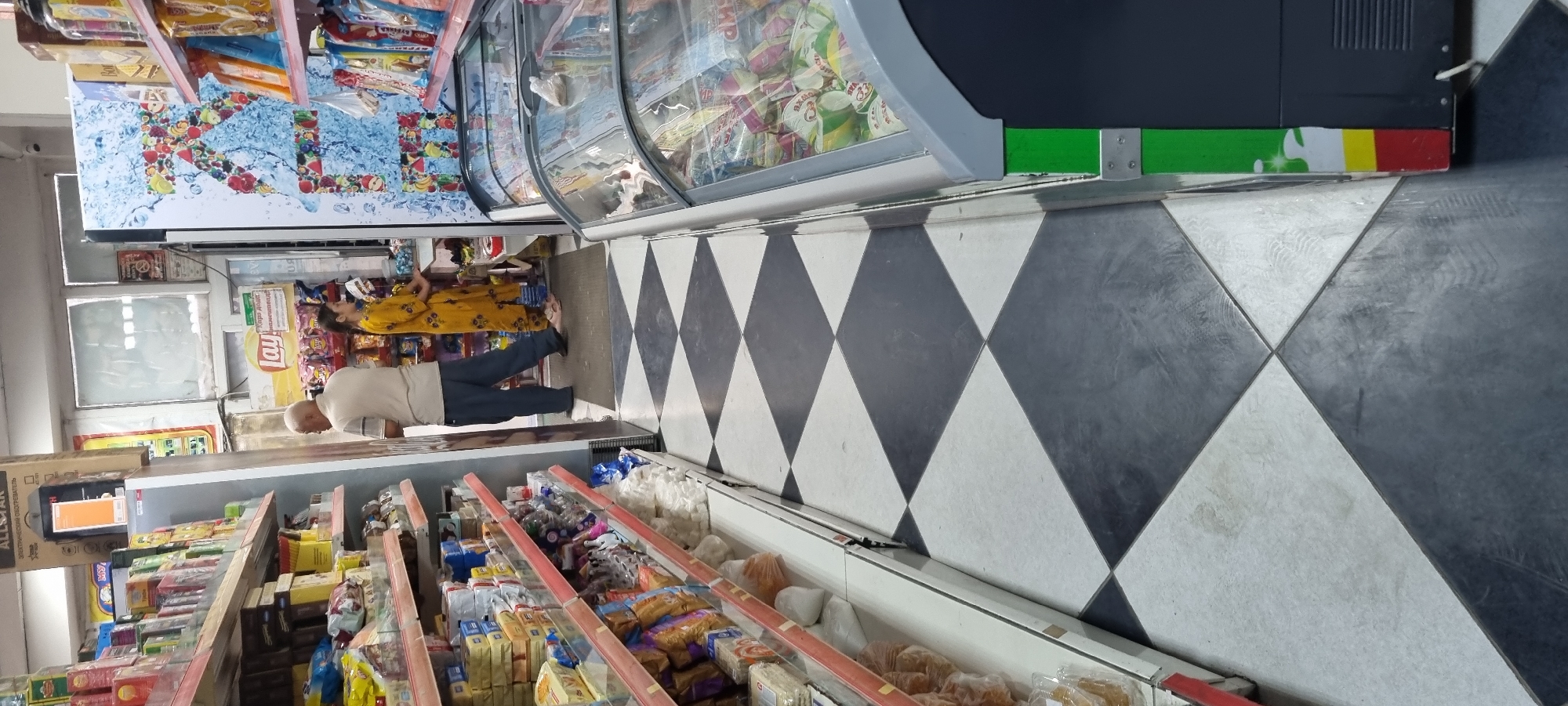 The image depicts the interior of a small grocery store, likely located in Ismoili Somoni, Tajikistan, based on the context provided. The flooring is a distinctive black and white diamond pattern. On the left, shelves are stocked with various food items, including packaged goods and snacks. To the right, a large display cooler with a colorful fruit and water design is visible. In the center aisle, an elderly man wearing a light-colored polo shirt and dark pants stands facing a younger woman dressed in a bright yellow, patterned outfit. They appear to be interacting near a checkout counter or display area. The overall impression is that of a busy, local convenience store.