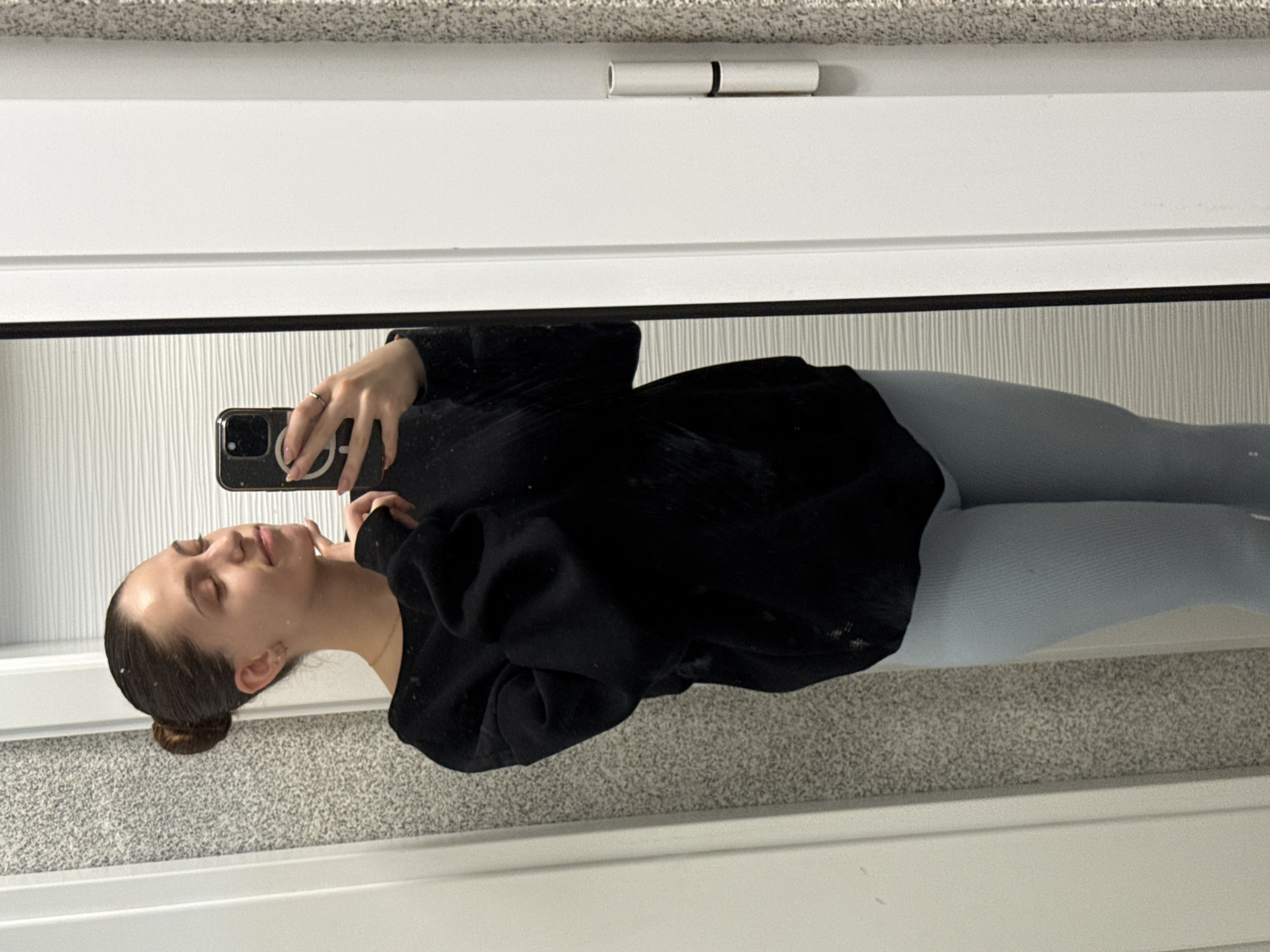 A female subject, appearing to be a young adult, with brown hair styled in a bun, is captured in a mirror selfie. She wears a black off-the-shoulder long-sleeved top and light blue ribbed leggings. Her eyes are closed, and a subtle smile is present. Her right hand, adorned with a gold-colored ring on the ring finger and manicured nails, holds a black smartphone with a visible white MagSafe ring, positioned to photograph her reflection. Her left hand's fingers touch her chin. Small gold-colored stud earrings are visible, along with a thin gold-colored necklace. The subject is reflected in a rectangular mirror with a thin black frame. To the viewer's left of the mirror, a light grey speckled or textured wall is visible. To the viewer's right of the mirror, a white door frame or panel is present, featuring two white cylindrical door hinges. The reflection in the mirror shows a wall with horizontal ridged patterns. The scene takes place indoors, in the city of Sofia, Bulgaria.