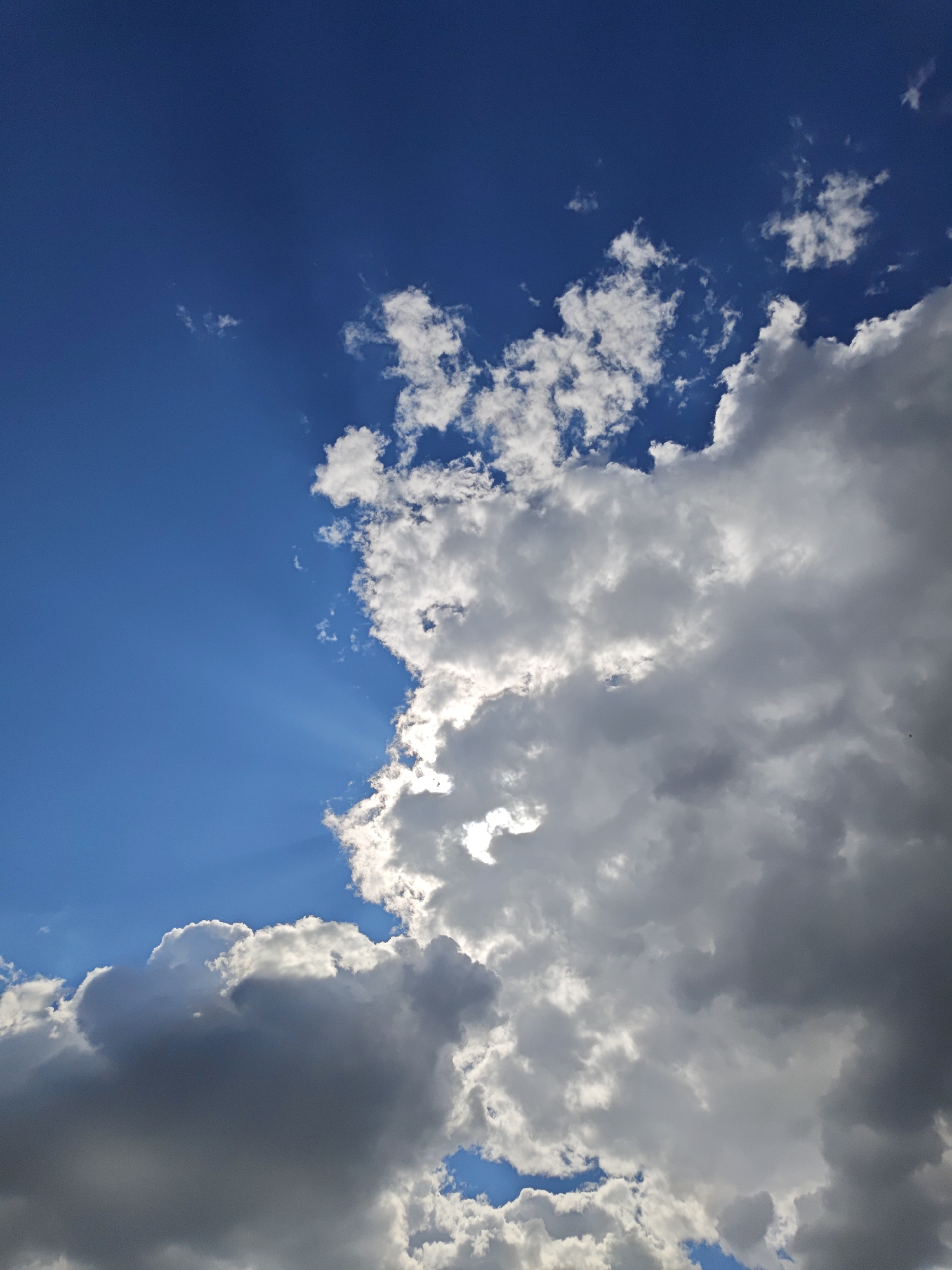 The image depicts a daytime sky, with a gradient of blue transitioning into a large mass of clouds. The upper left portion showcases a clear, deep blue expanse, fading slightly as it moves towards the center. This area is interspersed with small, scattered wisps of white clouds. The majority of the right side and lower portion of the image is dominated by large, voluminous clouds in shades of white and grey. These clouds appear to be dense, with some sections obscuring the sunlight, casting shadows within the cloud formation. Sun rays are visible as beams of light shining through gaps in the clouds. No specific location in Yekaterinburg, Russia can be determined from the image content. The scene primarily captures a dynamic interplay of light and shadow in the sky. There are no people, subjects, objects, actions, or interactions visible in the image apart from the natural elements.
