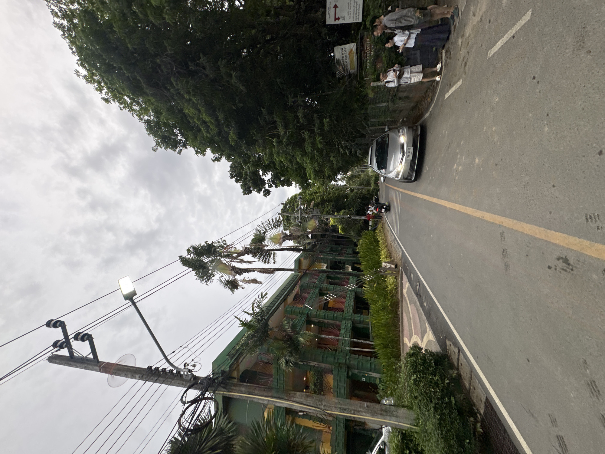This media file depicts a scene on a road in Karon, Thailand. The main subjects are a silver car driving on the road and several people standing on the sidewalk. A building with a distinctive green, multi-tiered facade and palm trees is visible on the left. A utility pole with many wires and a street lamp is also prominent on the left side of the frame.
The people appear to be standing and possibly conversing or waiting. The car is in motion. The building suggests a commercial establishment, possibly a hotel or restaurant, given the architecture and surrounding greenery. The presence of palm trees and lush vegetation indicates a tropical location.
The sky is overcast, suggesting it may be a cloudy day or early morning/late evening. There is no clear indication of a specific event taking place, but the scene captures a moment of everyday life on a street.
Visible text includes Thai script on signs near the people on the sidewalk, indicating directional information or business names. The car's license plate, "WD 4312," is also visible. The overall atmosphere is neutral, with no strong emotional cues conveyed. The lighting suggests it is not midday.