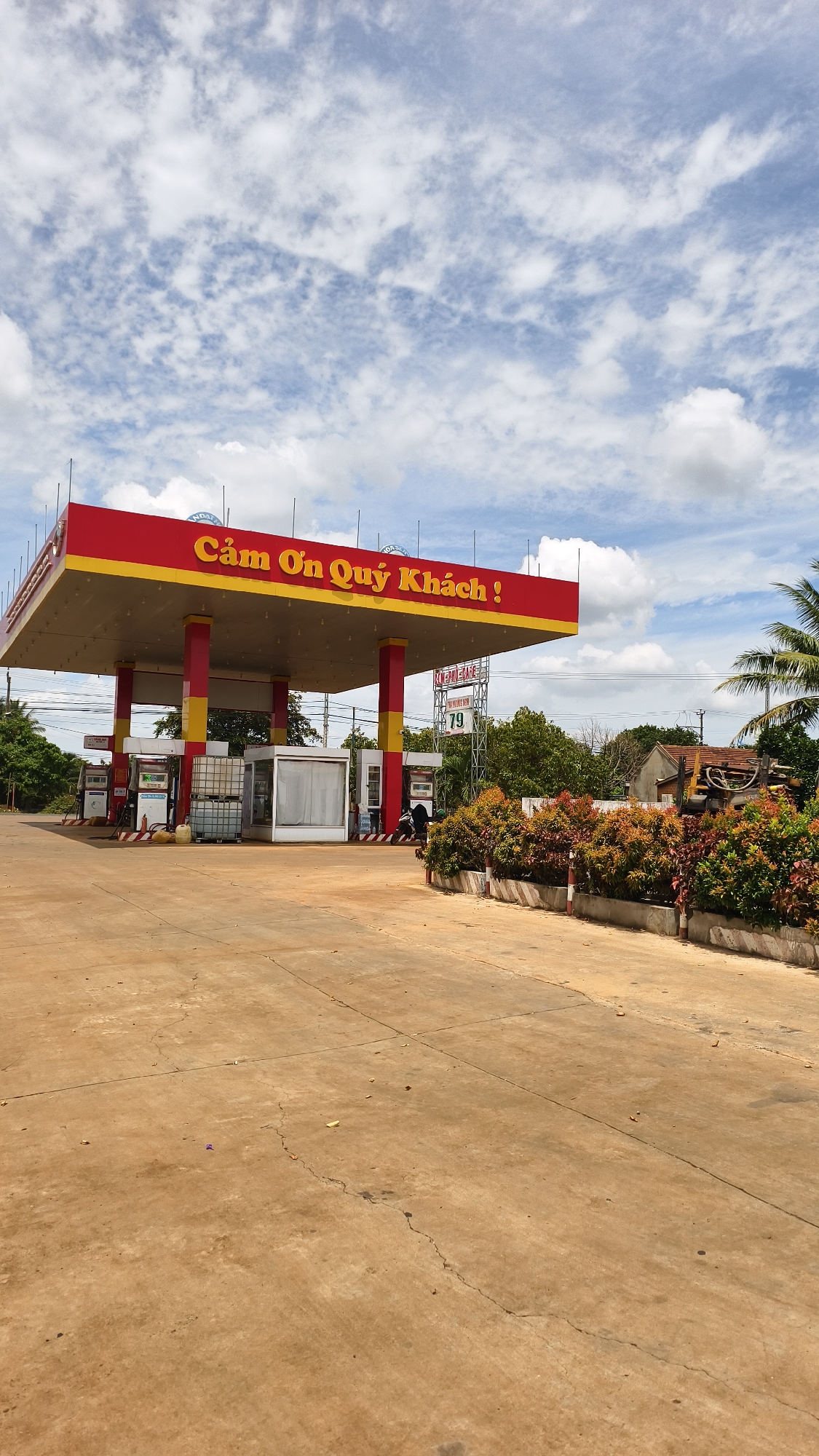This is a wide-angle, eye-level outdoor photograph of a gas station in Ia Hru, Vietnam. The gas station has a red and yellow canopy supported by red pillars. A sign above the canopy reads "Cảm Ơn Quý Khách!" in yellow lettering. Multiple gas pumps are visible under the canopy, along with a small white building. To the right of the gas station, there is a decorative border of red and green plants and a partial view of a building and trees. The ground is paved with concrete, showing some cracks and debris. The sky is blue with scattered white clouds. The scene is still and no human activity is explicitly depicted, though a motorcycle is partially visible to the right of the white building.
