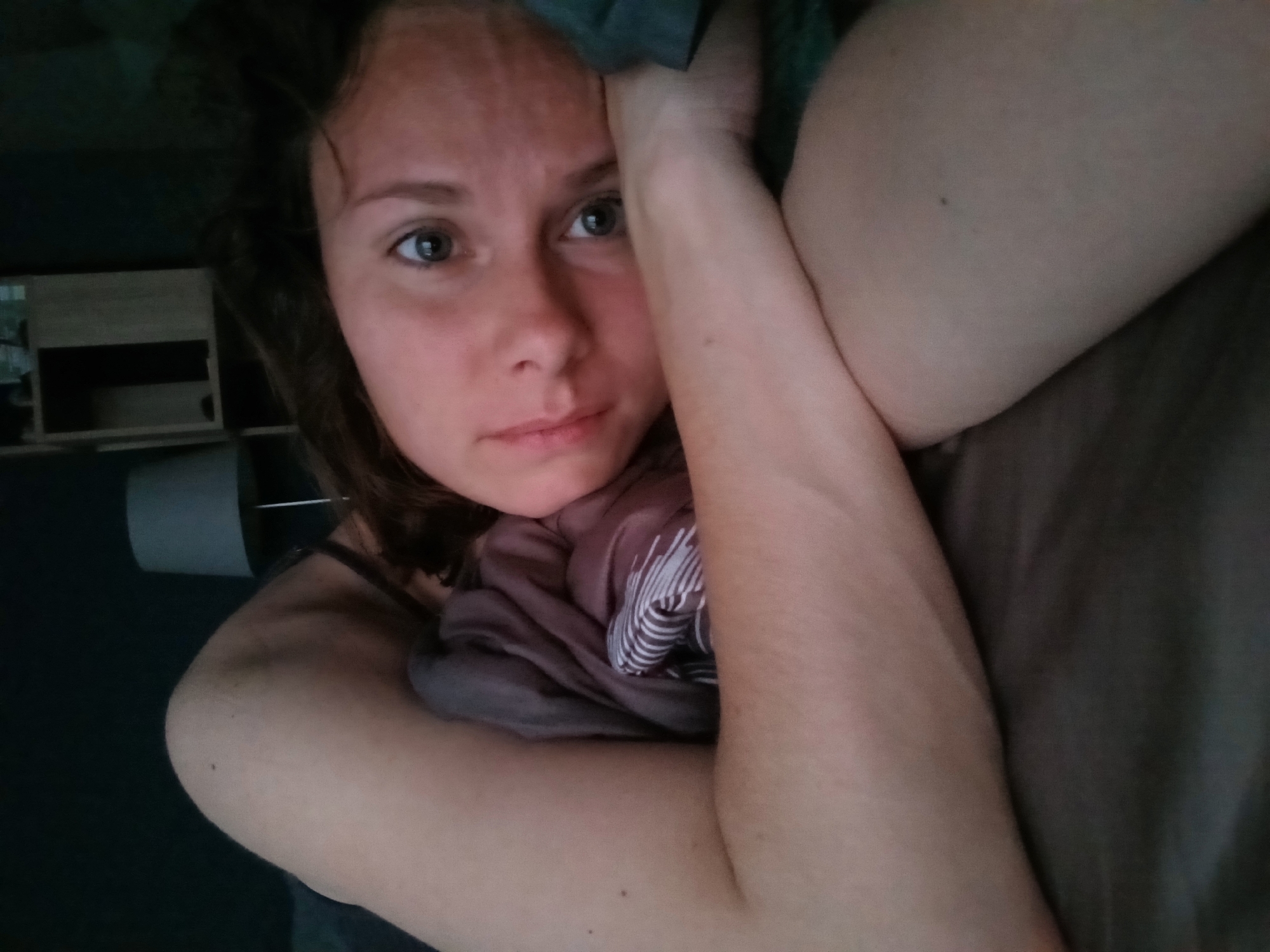 A young woman with brown hair and brown eyes is lying on a bed, looking towards the camera. She is partially covered by a purple and white striped blanket. The background is dark, with a lamp and part of a shelf visible. The image appears to have been taken in a bedroom.