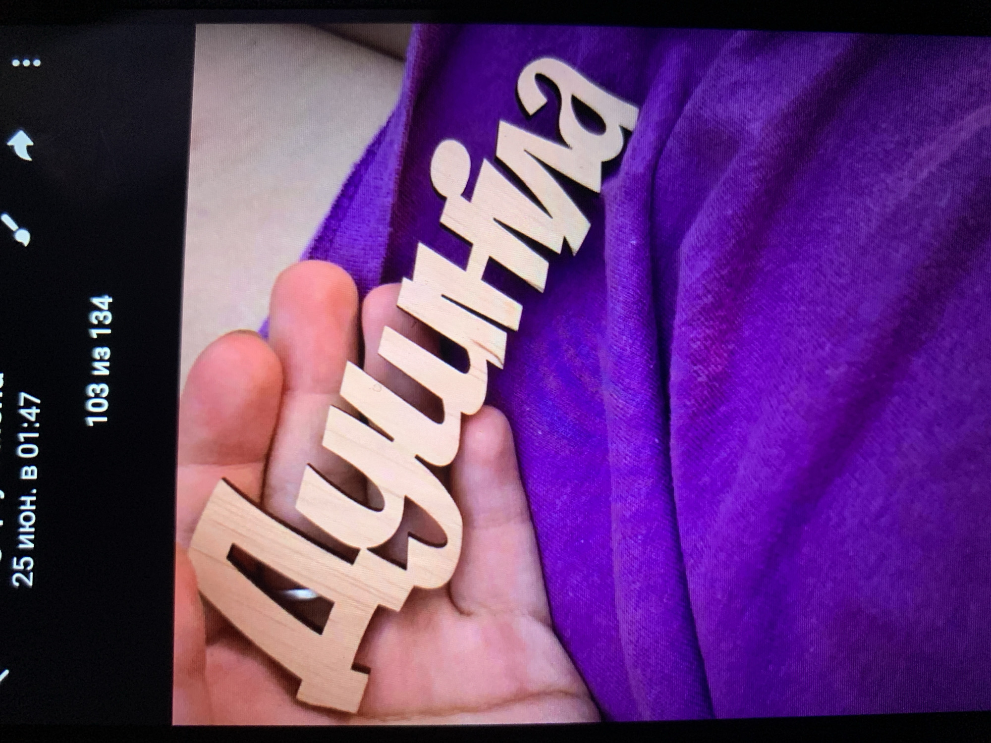 The image shows a close-up view of a wooden cutout of the name "Ауринка" (Aurinka) being held by a person's hand. The cutout is made of light-colored wood with a cursive font. The background consists of a wrinkled purple fabric. The top portion of the image displays text that appears to be from a digital device, showing "25 июня. в 01:47" and "103 из 134". There is no discernible information in the image or accompanying text to indicate any connection to the city of Lymanka, Ukraine.