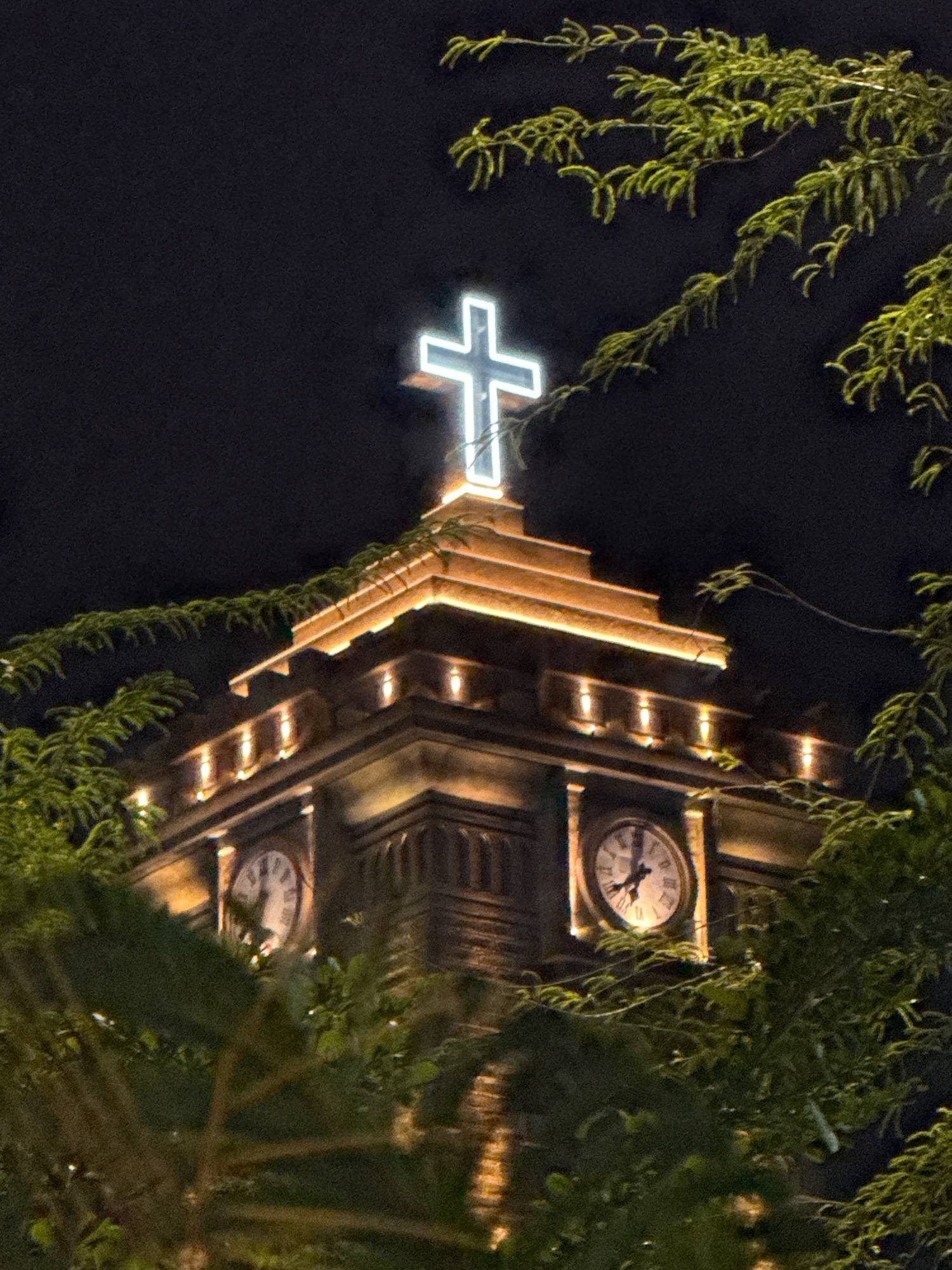The image captures a nighttime view of a tower in Nha Trang, Vietnam, adorned with a neon cross at its apex. The tower features two visible clock faces, one displaying Roman numerals. The structure is illuminated with warm-toned lights, highlighting its architectural details. Lush green foliage from surrounding trees partially obscures the tower, framing the scene. The background is a dark, featureless night sky. No people are visible in the frame.