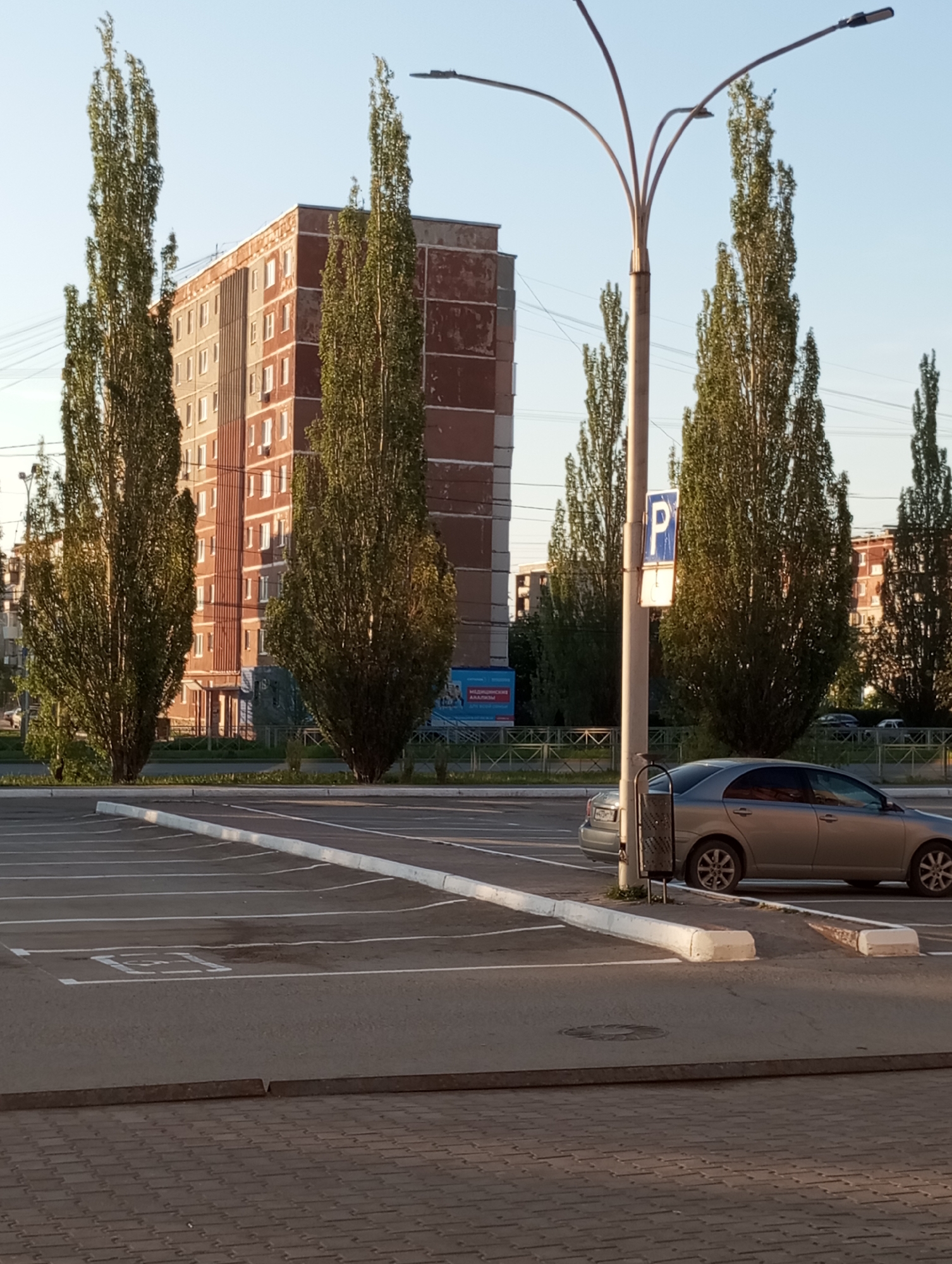 The image shows a parking lot in Kamensk-Uralsky, Russia. The parking lot is mostly empty with a silver car parked in one of the spaces. A parking sign is visible next to the car. In the background, there is an older apartment building with several tall, thin trees in front of it. A street lamp is positioned near the parked car. The parking lot is bordered by white curbs. The foreground shows a section of a paved area, possibly a sidewalk or pedestrian zone.