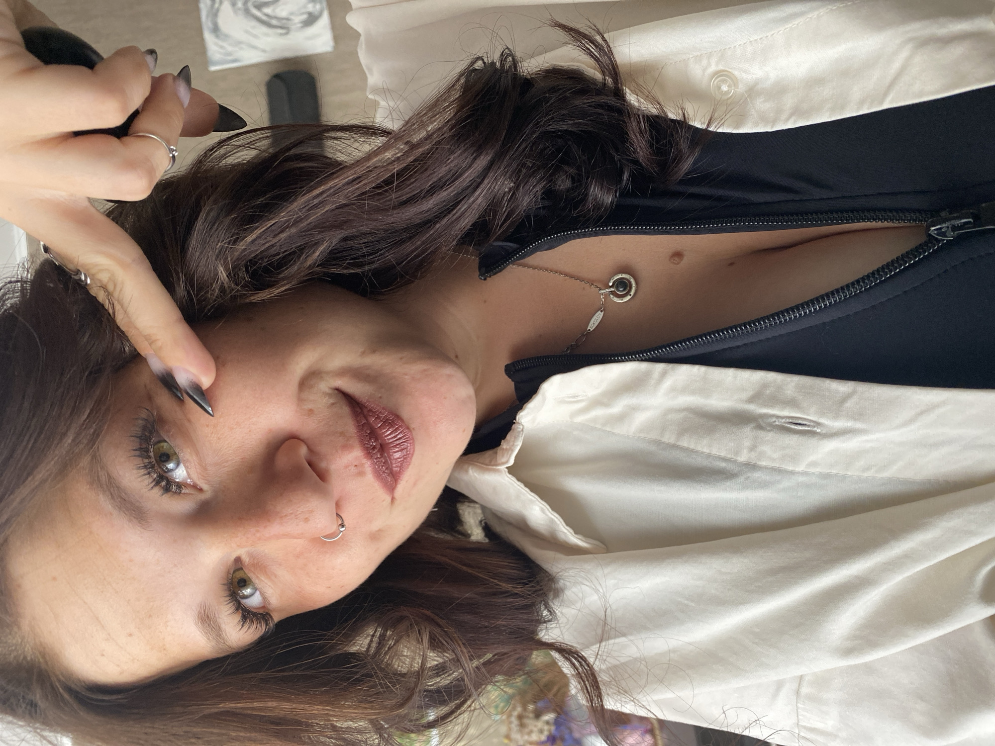 A woman with dark brown hair and green eyes is looking at the camera. She is wearing a white shirt over a black top and a necklace. Her right hand is raised, with her index finger pointing towards her left eye. She has long fingernails painted black. She is also wearing a nose ring and several rings on her fingers. The background is blurred and shows some indistinct objects. The image does not provide any information about the location in the city of Chaikovsky, Russia.