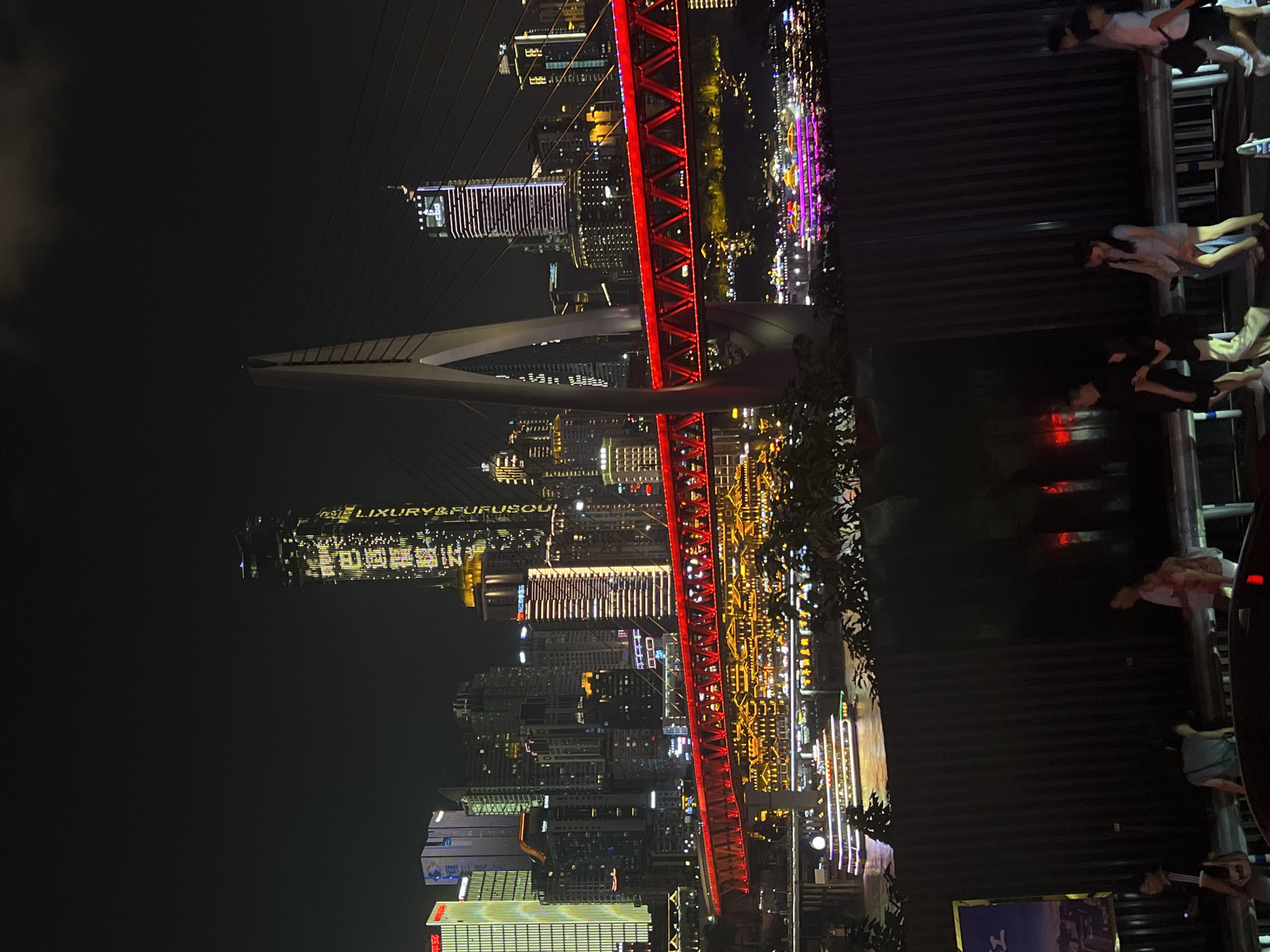 An nighttime, elevated, wide shot captures the cityscape of Jiangbeicheng Subdistrict, Chongqing, China. A modern suspension bridge with a prominent, pale gray pylon and red illuminated superstructure spans the frame horizontally across the mid-ground. Below the bridge, the river is visible, with a brightly lit, multi-deck passenger boat navigating the water. On the riverbanks, numerous buildings with illuminated windows and facades create a vibrant, urban panorama. In the far background, skyscrapers of varying heights and architectural styles are illuminated against the dark sky. In the foreground, a dark, corrugated metal barrier runs horizontally, obscuring the lower portion of the scene. Several individuals, appearing to be pedestrians, are dimly visible walking along a walkway behind the barrier. The overall scene is characterized by bright artificial lighting that accentuates the urban environment.
