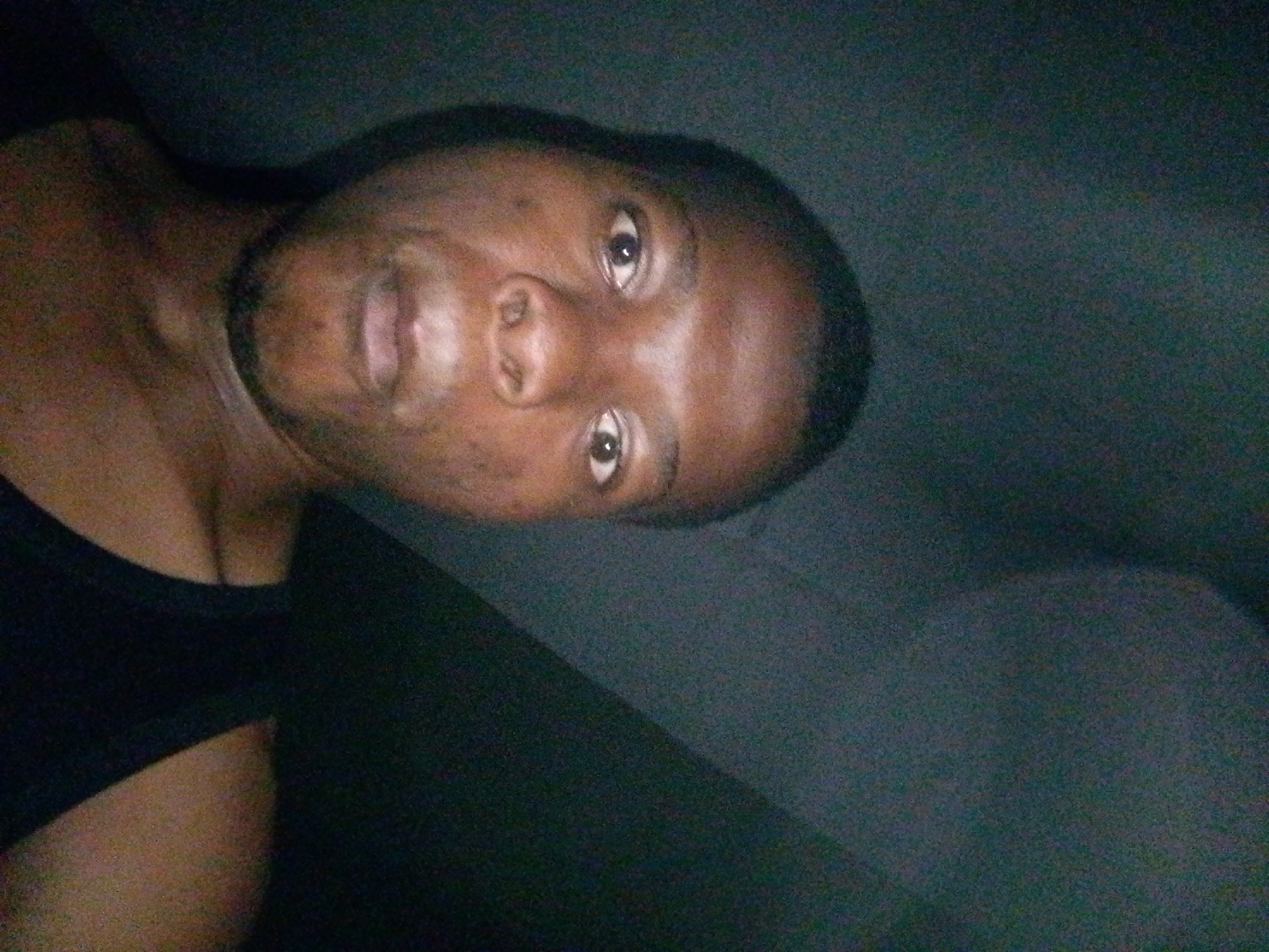 A close-up, dimly lit, and grainy selfie shows the face of a person with dark skin, looking directly at the camera. The image is rotated counterclockwise, so the person's head is tilted to the left. Their eyes are open, and their mouth is slightly ajar. They are wearing a black tank top, with the strap visible on the left side of the frame. The background is a dark, textured surface, possibly upholstery or fabric. The overall impression is of a casual, indoor setting. This image was taken in Ipetumodu, Nigeria.