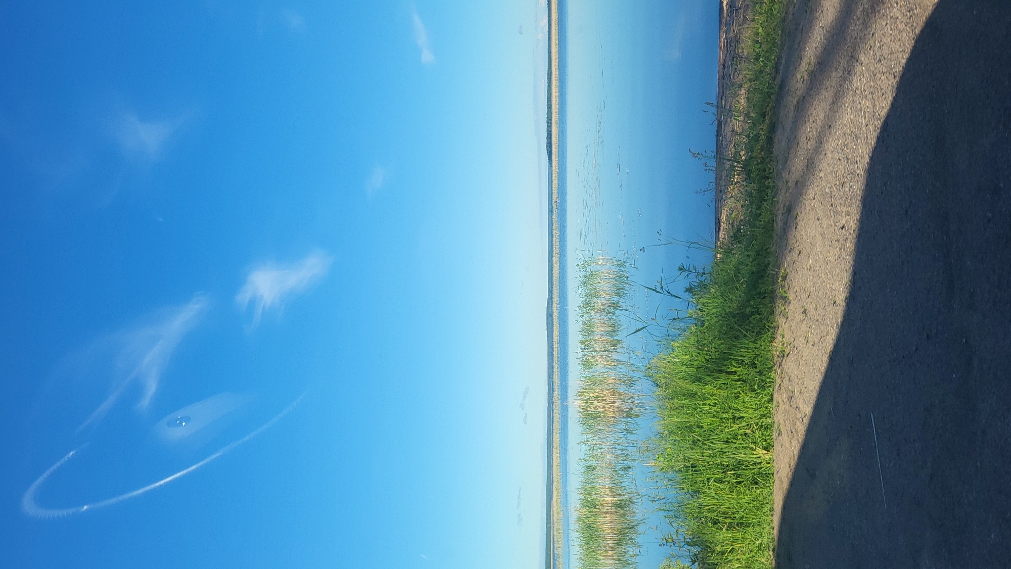 The media file displays a scenic landscape with a vast expanse of clear blue sky on the left, featuring a few wispy clouds and a circular contrail. The right side of the image is dominated by a tranquil body of water, likely a lake or a wide river, reflecting the blue sky and distant, hazy land formations. Along the shoreline, tall reeds and lush green grass are visible, with a dirt path or road bordering the grass on the far right. The sunlight suggests it is daytime, likely late afternoon or early evening given the long shadow cast by an unseen object on the road. The overall mood is serene and peaceful, evoking a sense of calm nature. There is no visible text within the image. The location context of Solnechnoe, Russia, suggests a possibly rural or coastal setting.