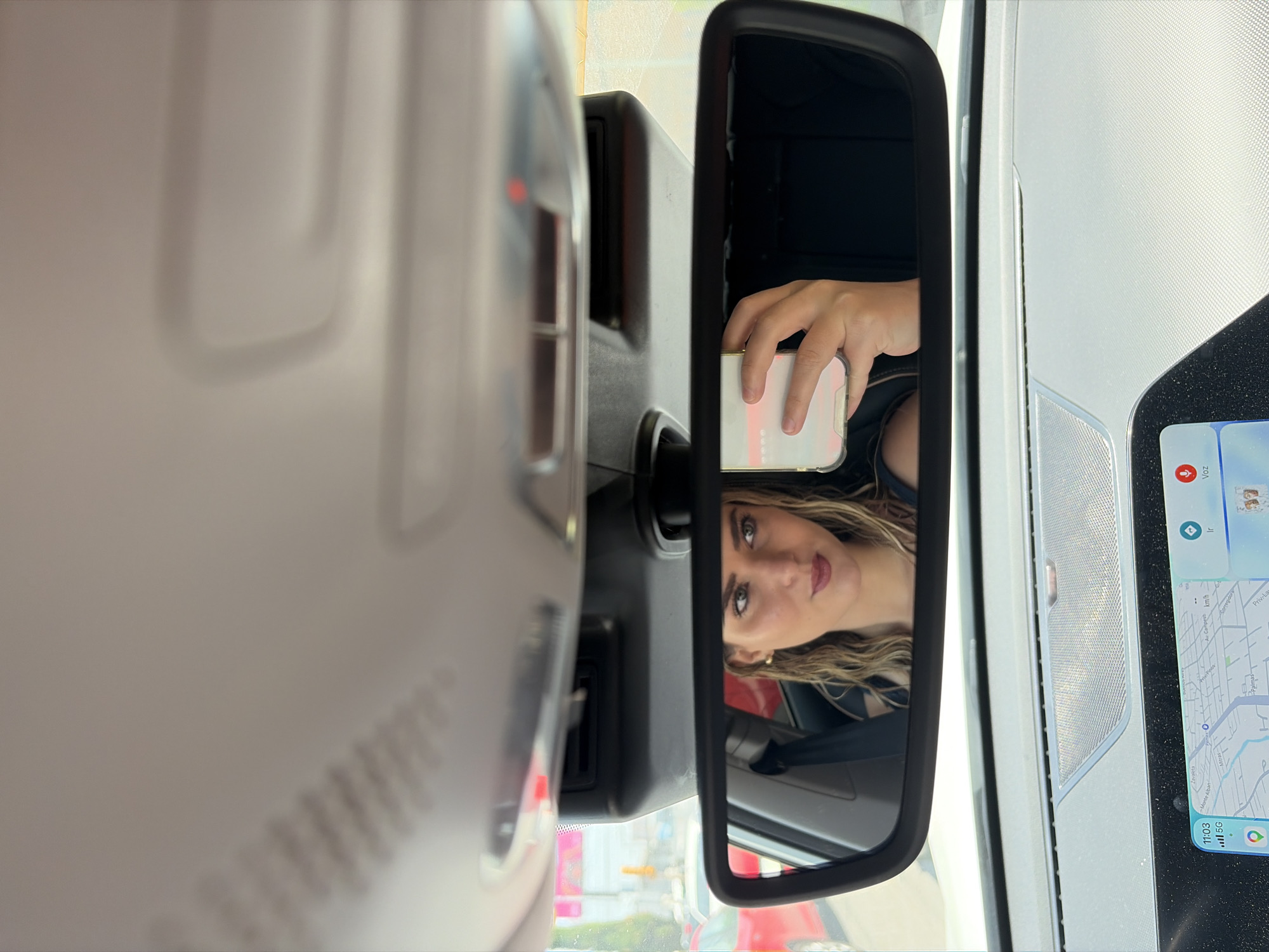 The image shows a woman's reflection in a car's rearview mirror. The woman is holding a smartphone in her hand, partially visible in the mirror. A car dashboard with a navigation screen is seen on the right side of the mirror, showing what appears to be a map of the city of San Bernardino Tlaxcalancingo, Mexico, as indicated by the street names visible on the screen. The time on the screen shows 11:03. The mirror is centrally located, with the interior roof of the vehicle visible above it, and reflections of external objects such as another car (possibly red), and street features can be seen in the bottom of the rearview mirror. The woman has medium-length wavy hair, light-colored eyes, and is wearing earrings. The image is taken from within the car, looking towards the rearview mirror.