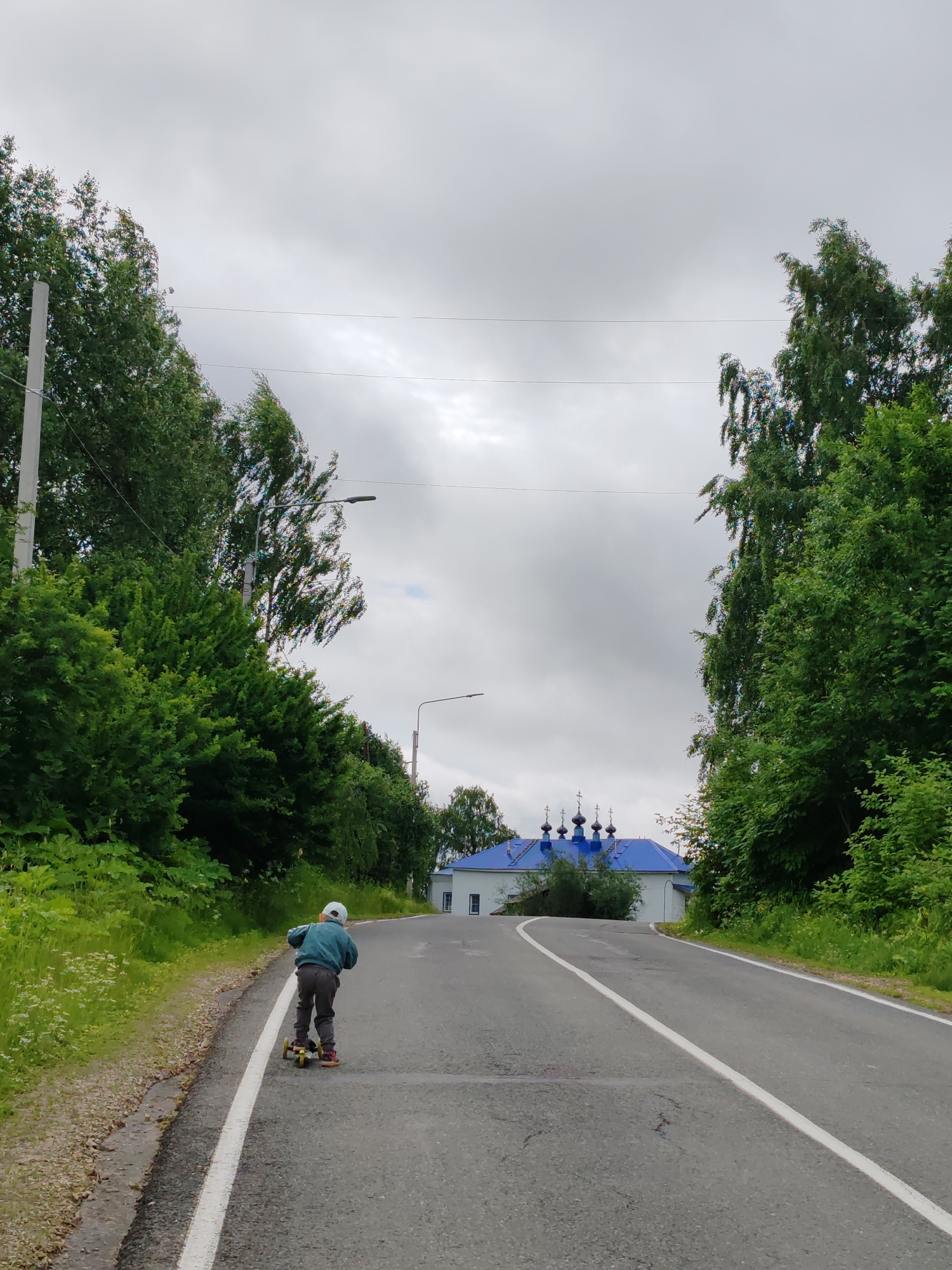 A young child wearing a light blue jacket, dark pants, and a white cap rides a yellow and black scooter on a paved road. The child is facing away from the camera, moving uphill. The road has a white dividing line. Lush green trees line both sides of the road. In the background, a white building with a bright blue roof and multiple domes, characteristic of a Russian Orthodox church, is visible. Power lines are suspended overhead. The sky is overcast with gray clouds. The scene appears to be set in the city of Chukhlem, Russia.