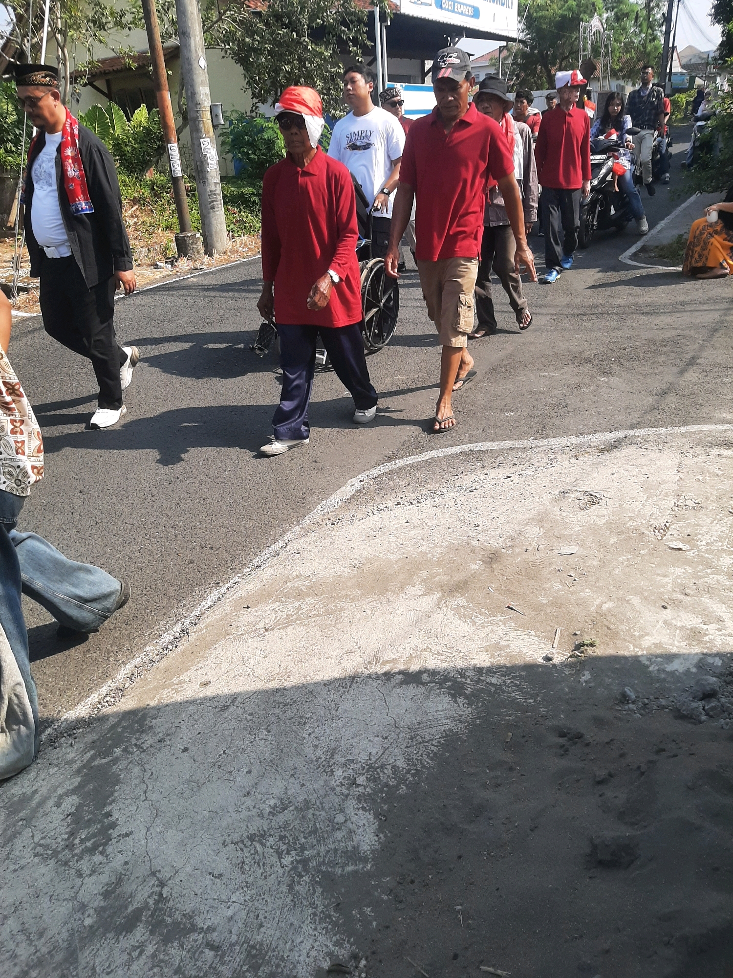In Sumampir, Indonesia, on a paved road under a bright sun, a group of people are walking. In the foreground, a person in a black jacket and white shirt, wearing a red and white scarf and a black cap, walks towards the left. To their right, another person in a red polo shirt and dark pants, wearing a red cap and sunglasses, walks with a wheelchair beside them. Further back, a man in a red polo shirt and tan shorts walks in flip-flops. Behind him, a woman rides a motorcycle. A man in a plaid shirt and dark pants walks in the background. The environment includes a sidewalk with vegetation and trees, as well as utility poles.