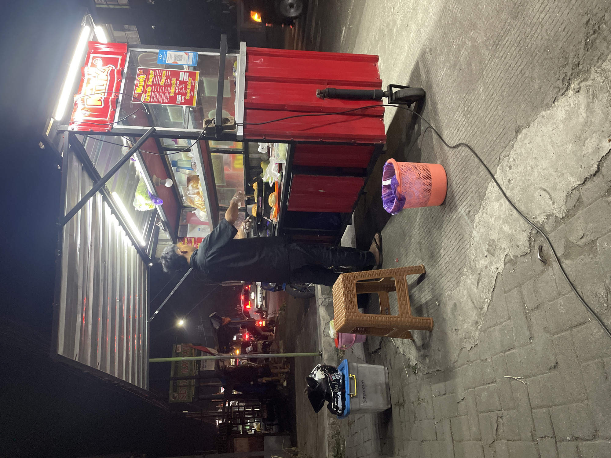 The image depicts a street food stall at night, likely in an urban environment. The stall itself is red and white, with a corrugated metal body and a metal frame supporting a canopy. Fluorescent lights illuminate the interior of the stall, showcasing food items and cooking equipment. A man in a black shirt and pants is standing behind the counter, preparing food. In front of the stall, a small orange plastic stool and a blue plastic bin with a helmet on top are visible. Further in the background, there are other stalls, vehicles, and pedestrians, suggesting a busy street. The ground is paved with interlocking concrete blocks and some sections of asphalt. A pink trash can with a purple trash bag is placed near the stall. The overall scene suggests a typical street food vending operation in a city at night.