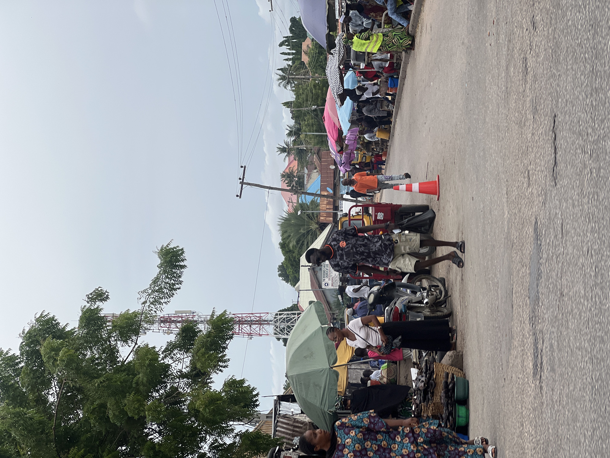 The image depicts a busy street scene, likely a marketplace, in Jalingo, Nigeria. A large red and white cell tower dominates the upper left portion of the image, partially obscured by a lush green tree. The street is paved with asphalt and appears worn in places.
Numerous people are present, engaged in various activities. In the foreground, a person wearing a vibrant, patterned dress walks towards the left side of the frame. To their right, a man in a grey and black patterned shirt and khaki shorts walks away from the camera. A motorcycle is parked near the center, with a person in a white shirt and black skirt standing next to it.
Further down the street, stalls are set up, indicated by colorful umbrellas providing shade. Several people are gathered around these stalls, some appearing to be vendors and others customers. A bright orange traffic cone is placed on the road in the middle ground. In the background, buildings with red roofs and palm trees are visible, suggesting a tropical or sub-tropical climate. The sky is overcast, indicating a cloudy day.
The overall impression is one of a bustling, active community market, typical of urban environments in Nigeria. The presence of the cell tower and the variety of clothing styles suggest a modern but distinctly Nigerian setting.