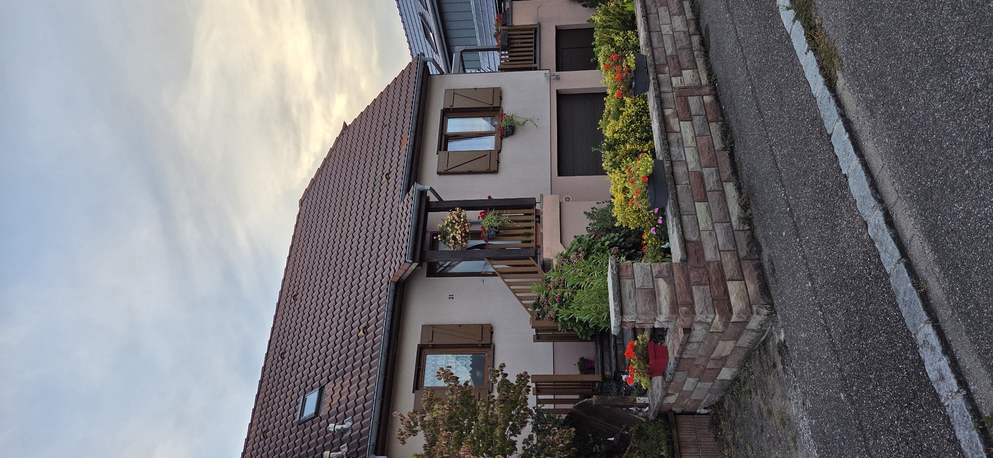 In Kilstett, France, a two-story house with a reddish-brown tile roof is visible. The house has a light pink facade, brown wooden shutters on the windows, and a small number "21" near the front door. A wooden staircase leads to the entrance, which is adorned with hanging flower baskets. In front of the house, a low wall made of multi-colored bricks surrounds a garden with various flowering plants and bushes, including yellow and red flowers in planters. A dark-colored garage door is present on the ground floor. The scene is captured on a cloudy day, with the sky showing a mix of blue and white hues. The foreground features an asphalt road with a stone curb.