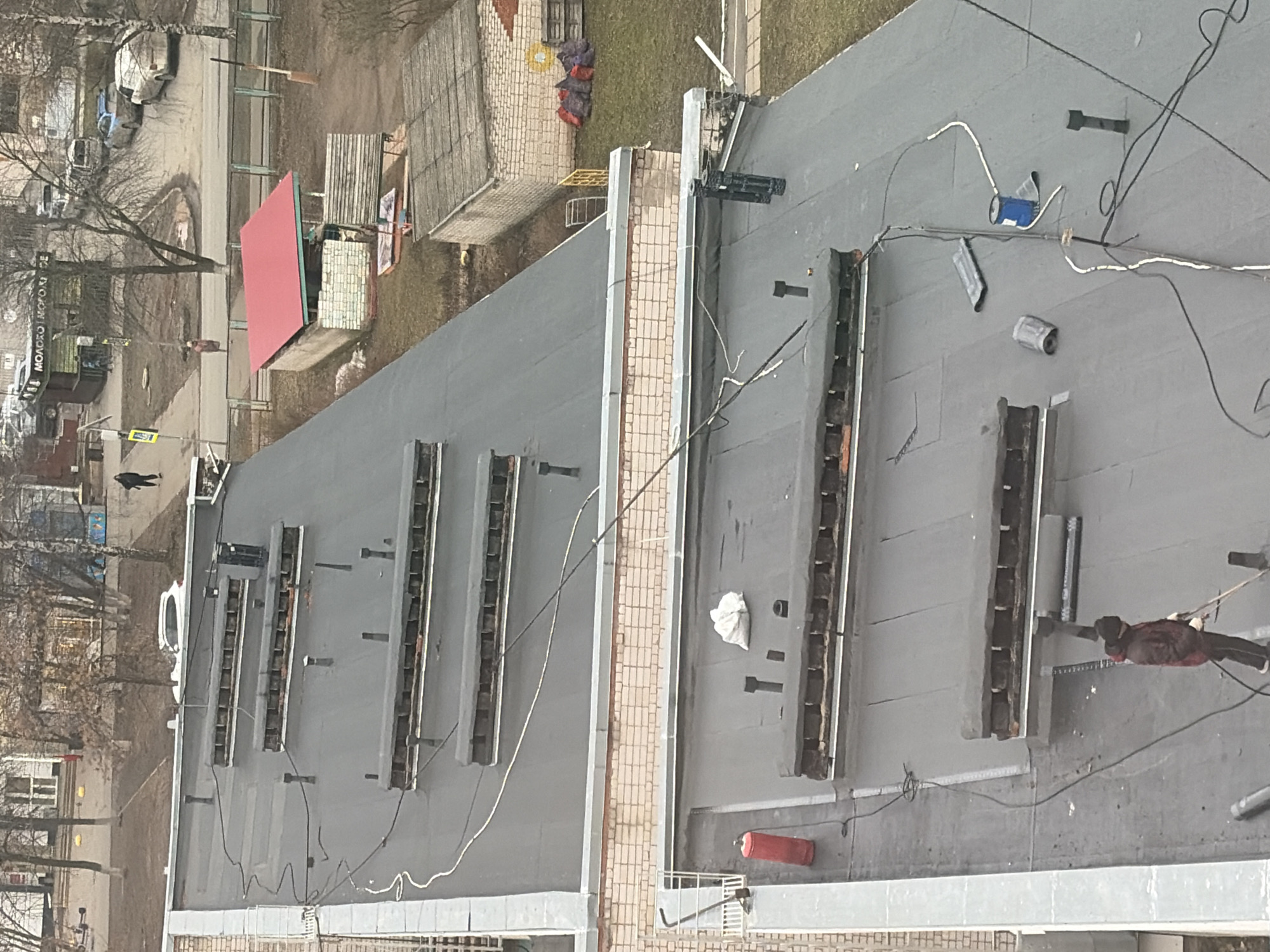 The image presents a high-angle view looking down from a building, capturing a flat rooftop in the foreground and a street scene beyond. The perspective is rotated, with the ground level appearing on the left and the rooftop on the right.
On the dark gray, flat roof, a person in a dark jacket is visible on the far right, seemingly engaged in work near one of several long, rectangular structures. These structures, possibly skylights or ventilation shafts, are arranged in rows and appear to be under construction or repair, showing exposed framing or internal components. Electrical cables, both black and white, are spread across the roof, connecting to various devices including a blue box, a metallic cylindrical object, and other tools. A red fire extinguisher and a white bag are also on the roof. A light-colored brick parapet separates the immediate roof area from the view below.
The ground level, visible beyond the rooftop, shows an urban environment. Bare-branched trees suggest a late winter or early spring season. A paved area or street features several parked cars and at least two people walking. A prominent red-roofed kiosk is visible, bearing a sign with Russian text: "MOLOKO | MOROZHENOE," which translates to "Milk | Ice Cream." Nearby are wooden pallets, construction materials, and a fence. The overall lighting is diffused, indicating an overcast daytime.
The scene depicts maintenance or construction activity on a building's roof, overlooking a typical urban street with daily pedestrian and vehicle traffic in Izhevsk, Russia.