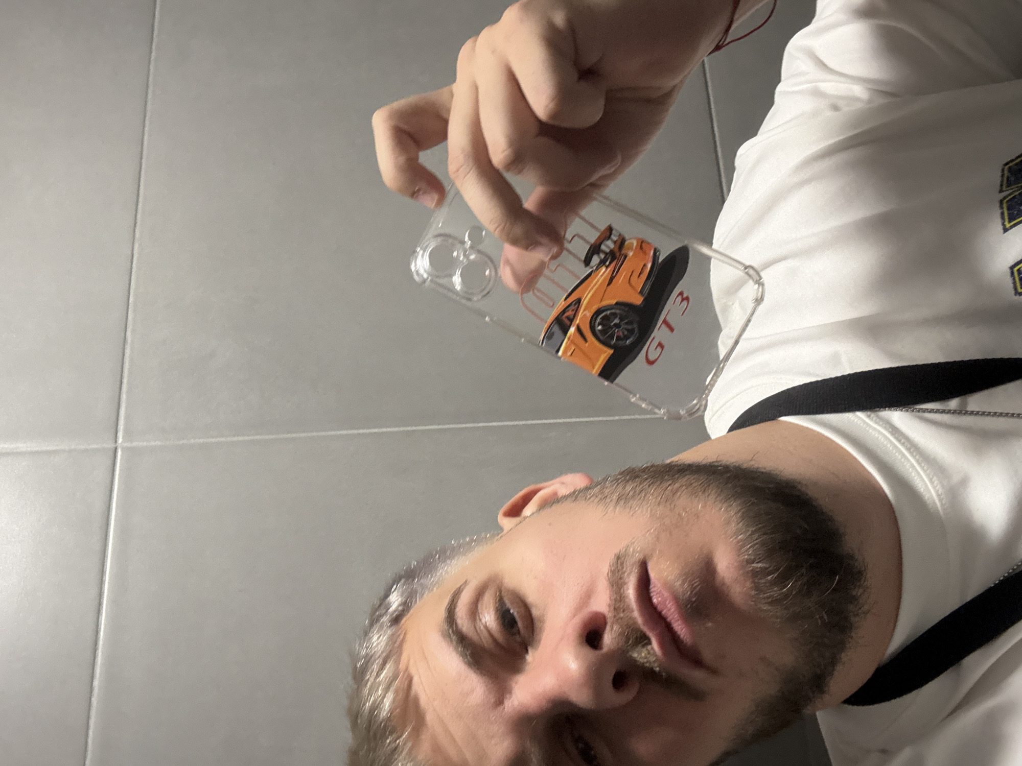 The image is a selfie taken from a low angle. A Caucasian male with salt and pepper hair, a beard, and gray eyes fills the lower portion of the frame. He is wearing a white T-shirt with black accents. The male is holding a clear phone case in his hand that features an orange sports car, labeled "GT3." The background is a light gray tiled wall. No information is available to determine the location in Sofia, Bulgaria.