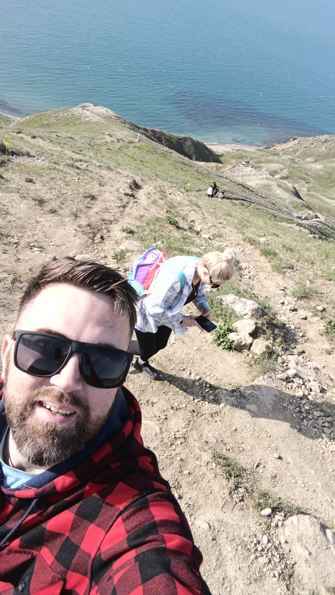 A selfie photograph features a man in the foreground wearing sunglasses and a red and black checkered hooded sweatshirt, with a blurred background of a woman holding a phone on a hillside overlooking a sea. The woman has blonde hair pulled back in a bun and is wearing a light-colored plaid shirt and a backpack. Two additional figures are visible further down the hillside. The terrain is primarily dirt and sparse vegetation. The location is Unknown, Unknown.