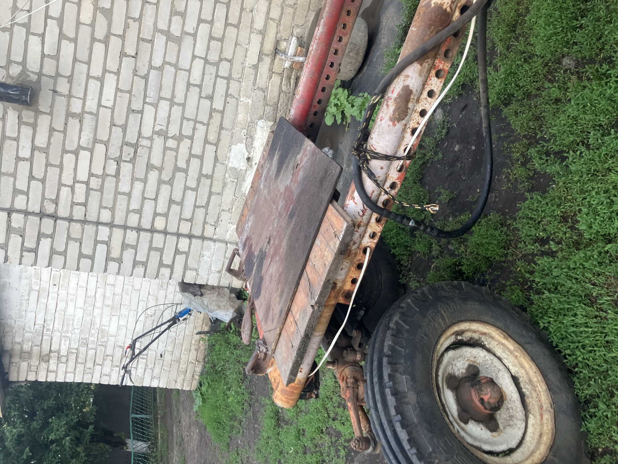 In the city of Vovkivka, Ukraine, a rusted, two-wheeled tractor with a wooden platform attached is parked on overgrown grass next to a white brick building. The tractor has a single large tire on the left side and a smaller wheel assembly on the right, also partially obscured by vegetation. A black hose is attached to the tractor and runs along its frame. In the background, a section of a green metal fence and a tree are visible.
