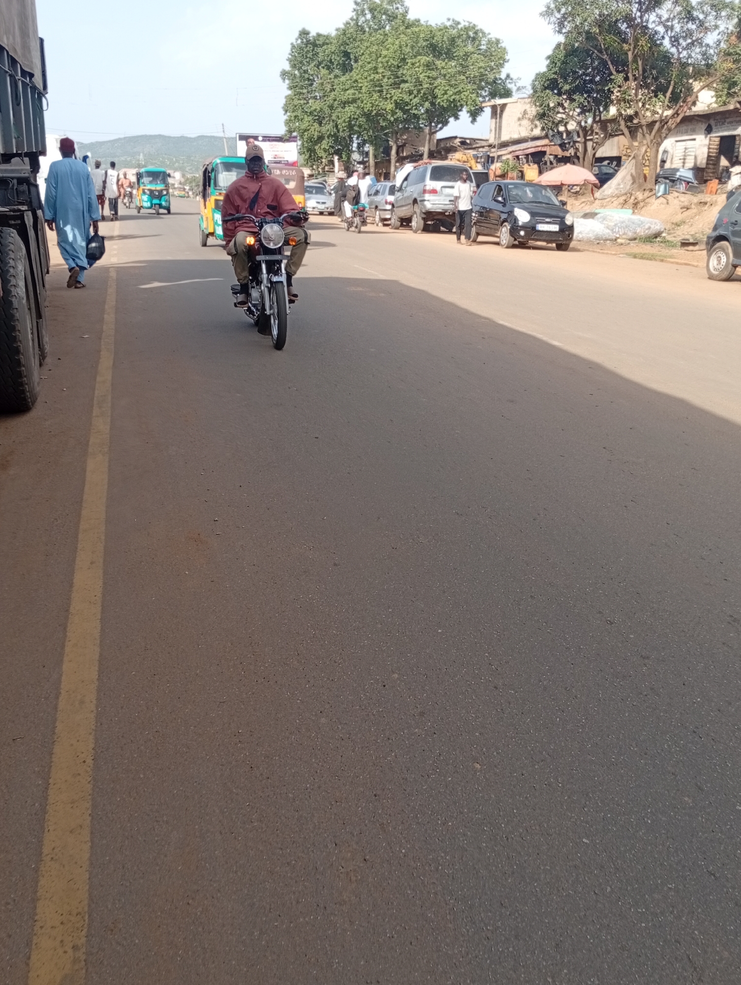 This is a scene from the city of Rafin Par, Nigeria, showing a street with several vehicles and people. On the left, a large truck is partially visible, with its tires and side taking up a significant portion of the frame. A man in a blue traditional robe and headscarf is walking away from the truck, carrying a dark bag. Further down the street, a man wearing a brown jacket and a dark cap is riding a motorcycle. He is wearing dark pants and dark shoes. Behind him, a bright yellow and green auto-rickshaw is visible. Several other vehicles, including a black car, a silver SUV, and another white car, are parked or moving on the right side of the road. Pedestrians can be seen walking on the side of the street. The environment appears to be a typical urban street with buildings and trees lining the road. The asphalt road is marked with a yellow line on the left side. The sky is bright and appears to be clear, suggesting daytime.