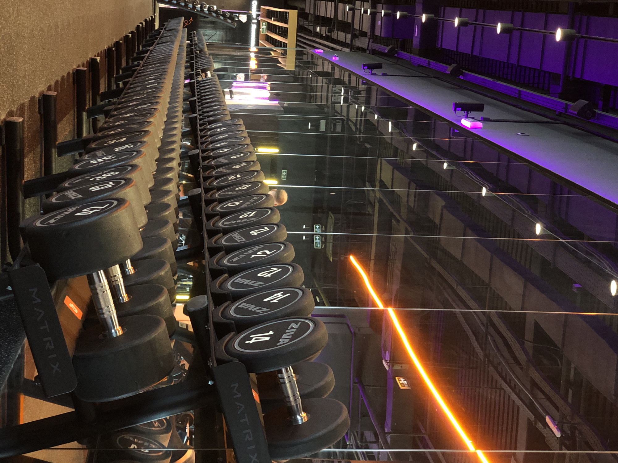 A modern gym interior in Krasnoyarsk, Russia, is captured from a low angle, showcasing long rows of dumbbells on black metal racks. The image is oriented vertically, with the rows of weights extending into the background on the left and right. The dumbbells are black with silver handles, featuring white numbers indicating their weight, such as "12", "14", "24", "26", "28", "30", "32", "34", "36", and "38". The brand "ZIVA" is visible on many of the dumbbell ends, and the word "MATRIX" is seen vertically printed on the side of the dumbbell racks.
The floor is highly reflective, acting like a mirror and creating a strong sense of depth by mirroring the dumbbells, overhead structures, and lighting. A prominent, bright orange/amber strip of light cuts diagonally across the reflective floor. Other lighting includes purple ambient lights and various white or yellowish spot lights, some of which are reflected on the floor and ceiling-like surfaces.
In the reflections, the head of at least one person is discernible, along with what appear to be overhead structures and other gym equipment. Green "Exit" signs with arrows are visible in the reflections on the far side of the gym. The overall atmosphere is sleek and contemporary, illuminated by the distinctive artificial lighting.