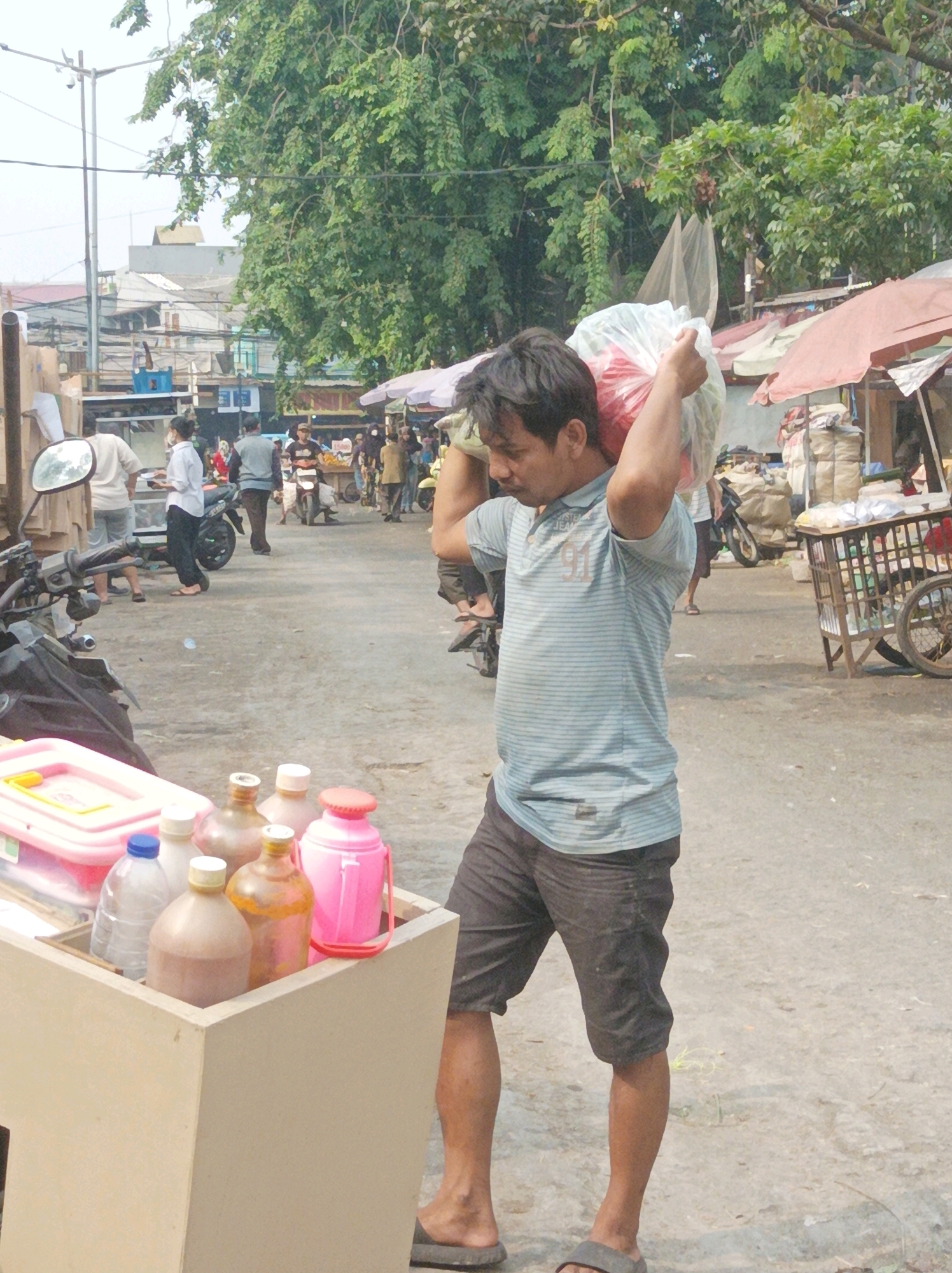 An Indonesian man carries a large plastic bag of goods over his shoulder in an outdoor market setting. He is wearing a light blue and white horizontally striped t-shirt and dark shorts. In the foreground, to the left, is a wooden cart filled with various bottles and a pink plastic container. Behind the man, to the right, is a wheeled cart with goods and various stalls with awnings. Other people and motorcycles are visible in the background. The scene appears to be a busy marketplace in South Jakarta, Indonesia, with a focus on commerce and street vending. The ground is unpaved and dusty, and trees and buildings line the street.