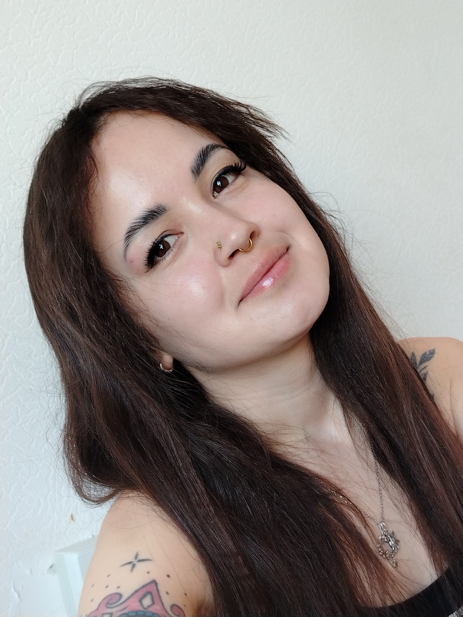 A woman with long, dark brown hair poses for a close-up photograph against a light-colored wall. She has fair skin, brown eyes, and a gold nose ring. The woman wears a necklace and earrings. Tattoos are visible on her shoulder and arm. The location of the photograph is not evident from the image. There is no indication that the location is in the city of Noviy, Russia.