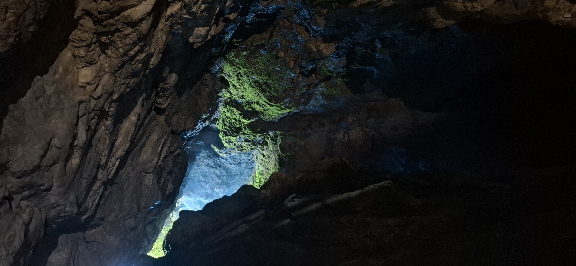 The image depicts the interior of the Devil's Throat Cave, located near the village of Trigrad, Bulgaria. The cave's ceiling is rugged and appears to be made of rock formations. A bright opening is visible in the upper left section of the cave, allowing natural light to filter in, creating a stark contrast with the darker interior. Lush green moss covers some of the rock surfaces, particularly on the right side of the opening. The lower portion of the cave is enveloped in deep shadow, with hints of blue light suggesting the presence of water or reflective surfaces in the darkness. There are no people or active events captured in the photograph; it is a static view of the cave's geological features.